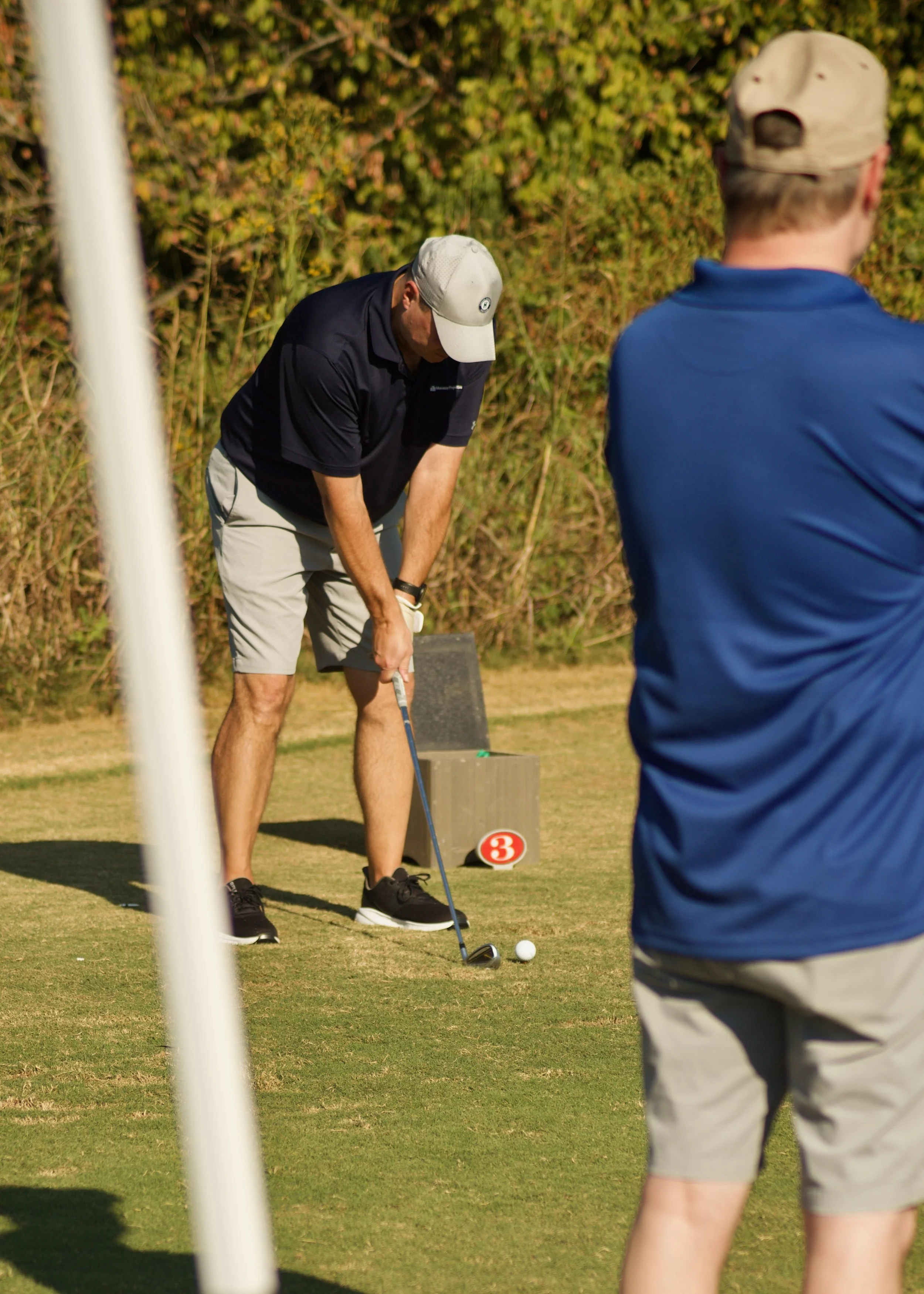 Two men playing golf on the course, one about to hit the ball, with a tree-lined background and a small numbered marker in the grass.