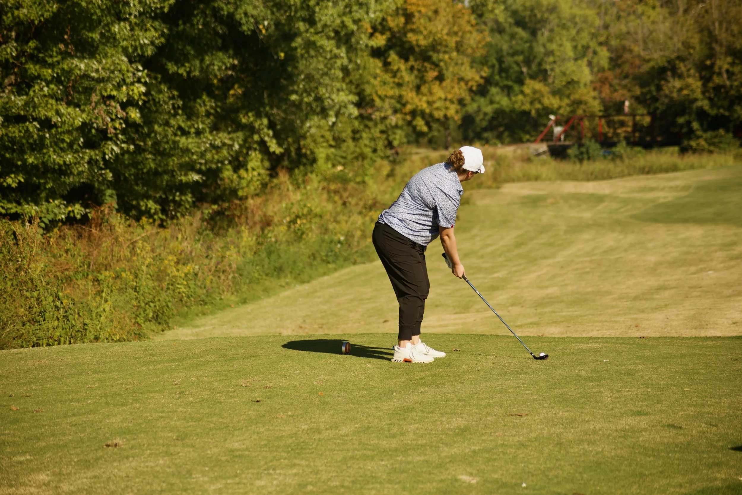 A woman with a white cap and gray shirt preparing to hit a golf ball on the green, surrounded by trees and a small bridge in the background.