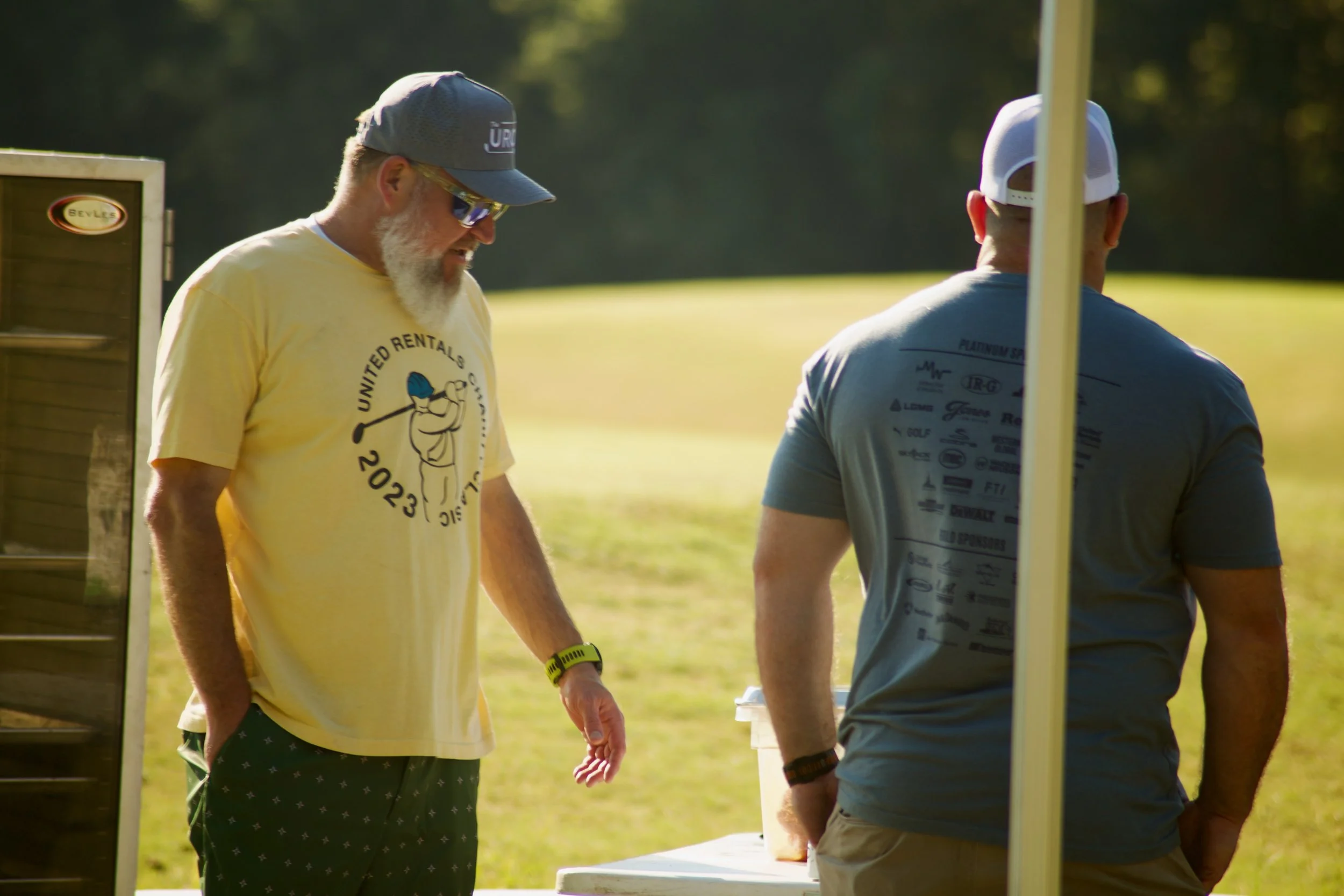 Two men engaged in conversation outdoors on a sunny day, standing near a table with drinks. One man is wearing a yellow t-shirt with a golf-themed logo, a gray cap, sunglasses, and a yellow wristband. The other man is in a gray t-shirt and a white ca