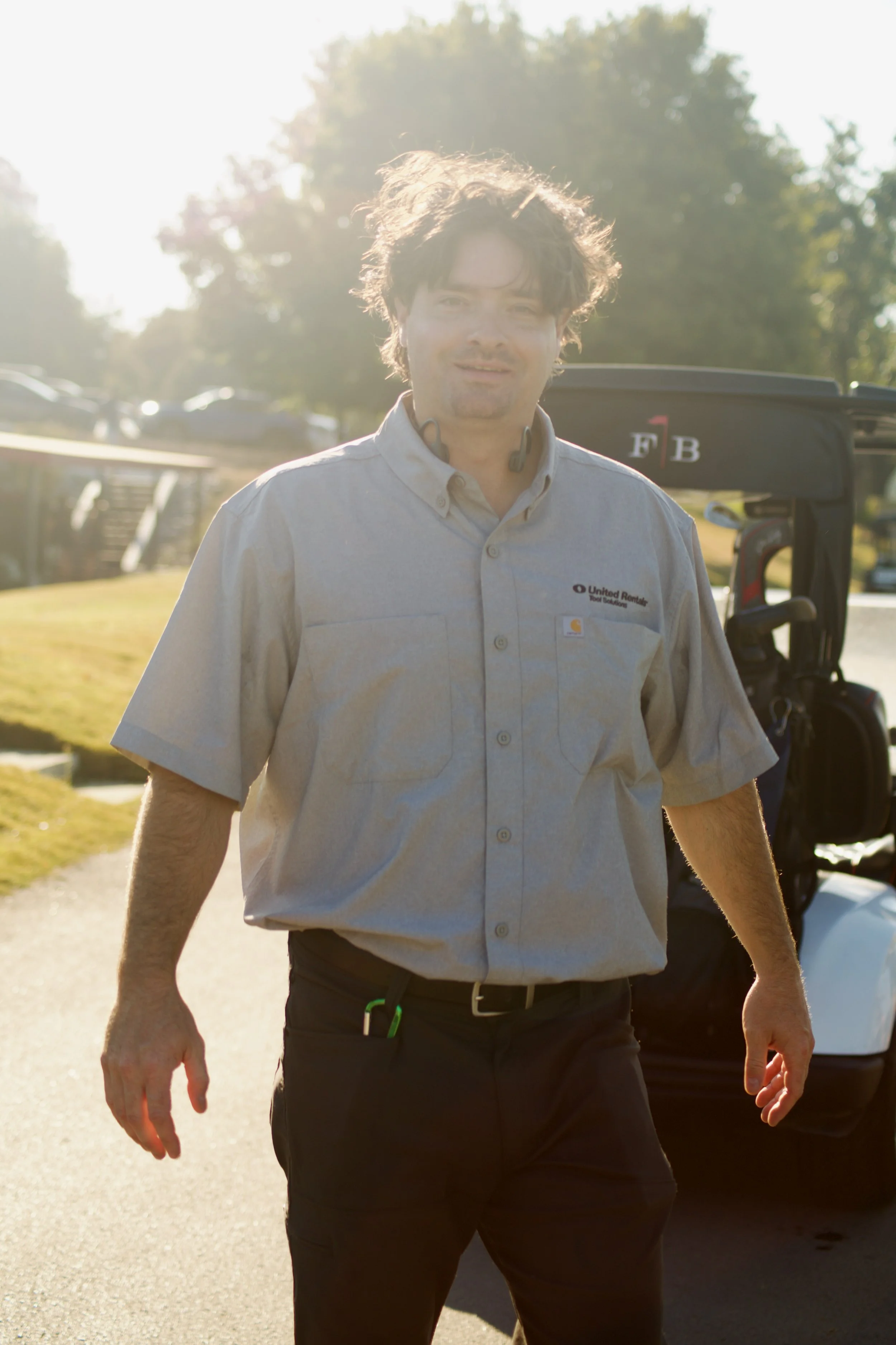 A man in a beige work shirt with a logo that says 'United Rentals' on the chest, standing outdoors during daylight, with trees and cars in the background. The sun is bright and shining toward the camera, creating a backlit effect.