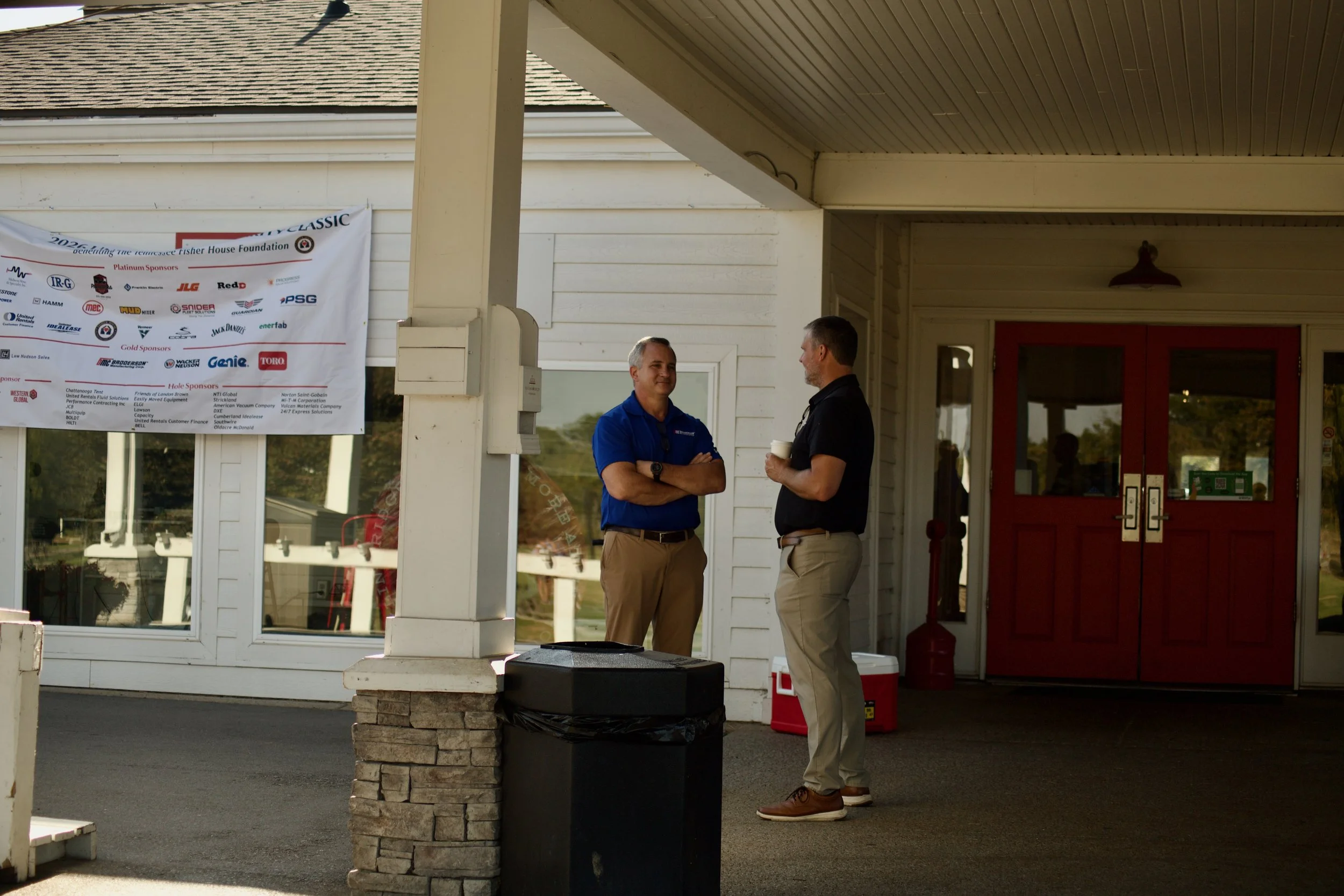Two men stand outside a building, engaged in conversation. One wears a blue shirt and khakis, the other a black shirt and tan pants, holding a cup. A banner with multiple logos hangs on the wall behind them, and the building has red double doors.