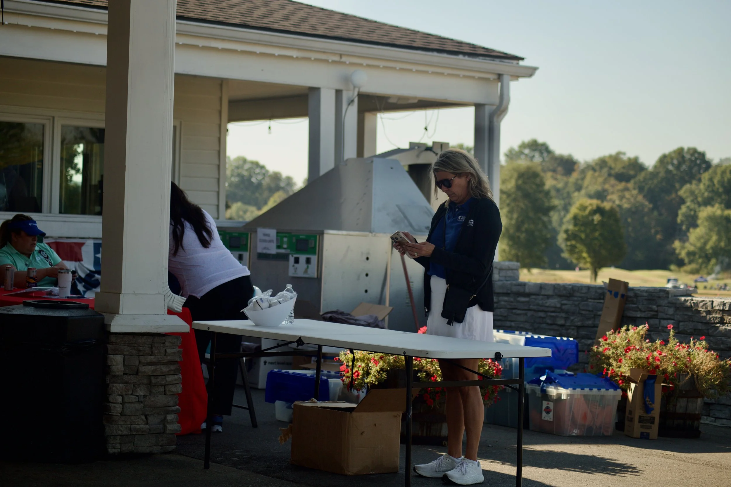 People at an outdoor event, with a woman in sunglasses looking at her phone, surrounded by tables with items, boxes, and flowers, under a covered patio on a sunny day.