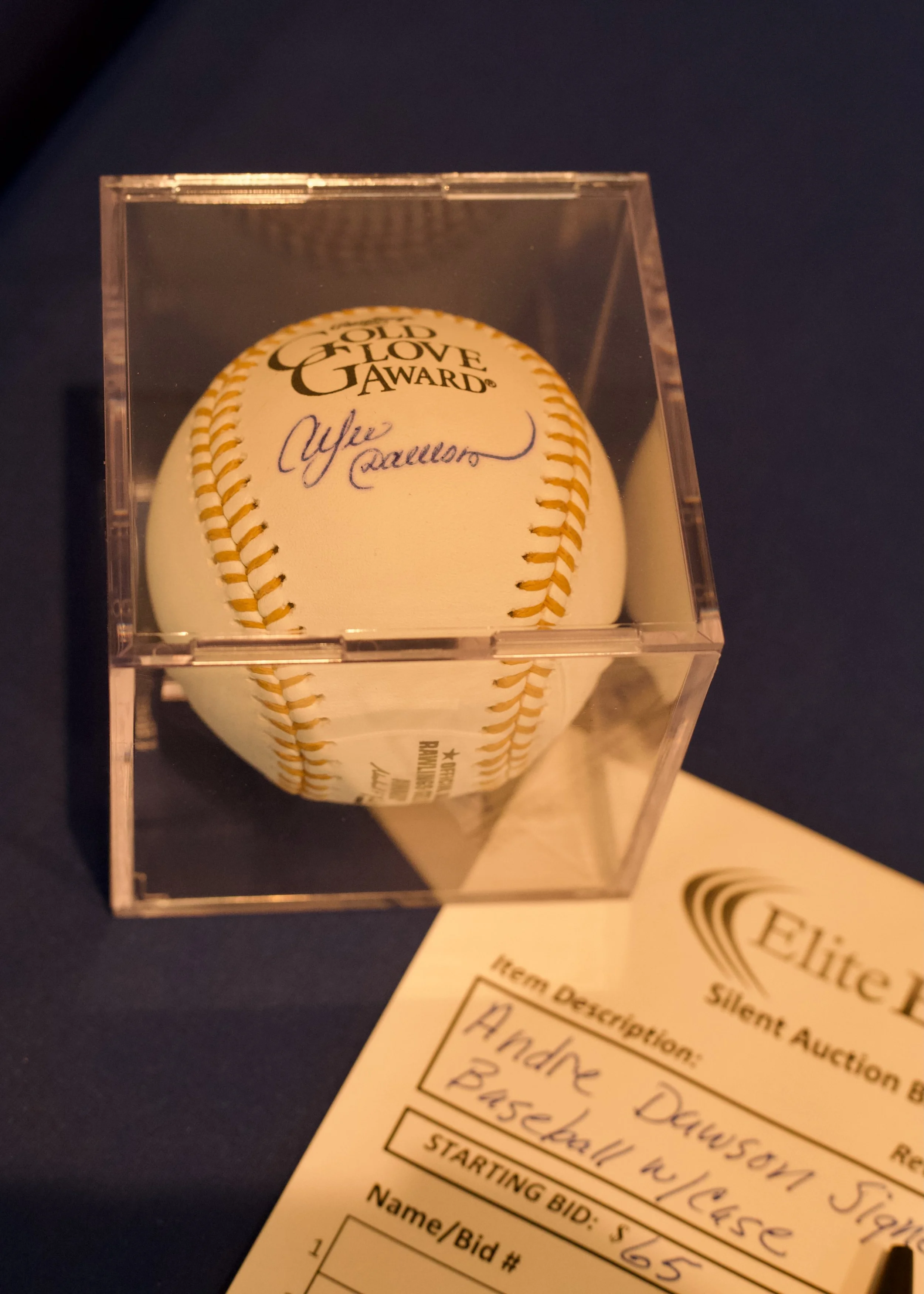A signed baseball inside a clear display case with a sticker labeled 'Old Love Gawrd' on top, displayed on a dark surface next to an auction sheet.