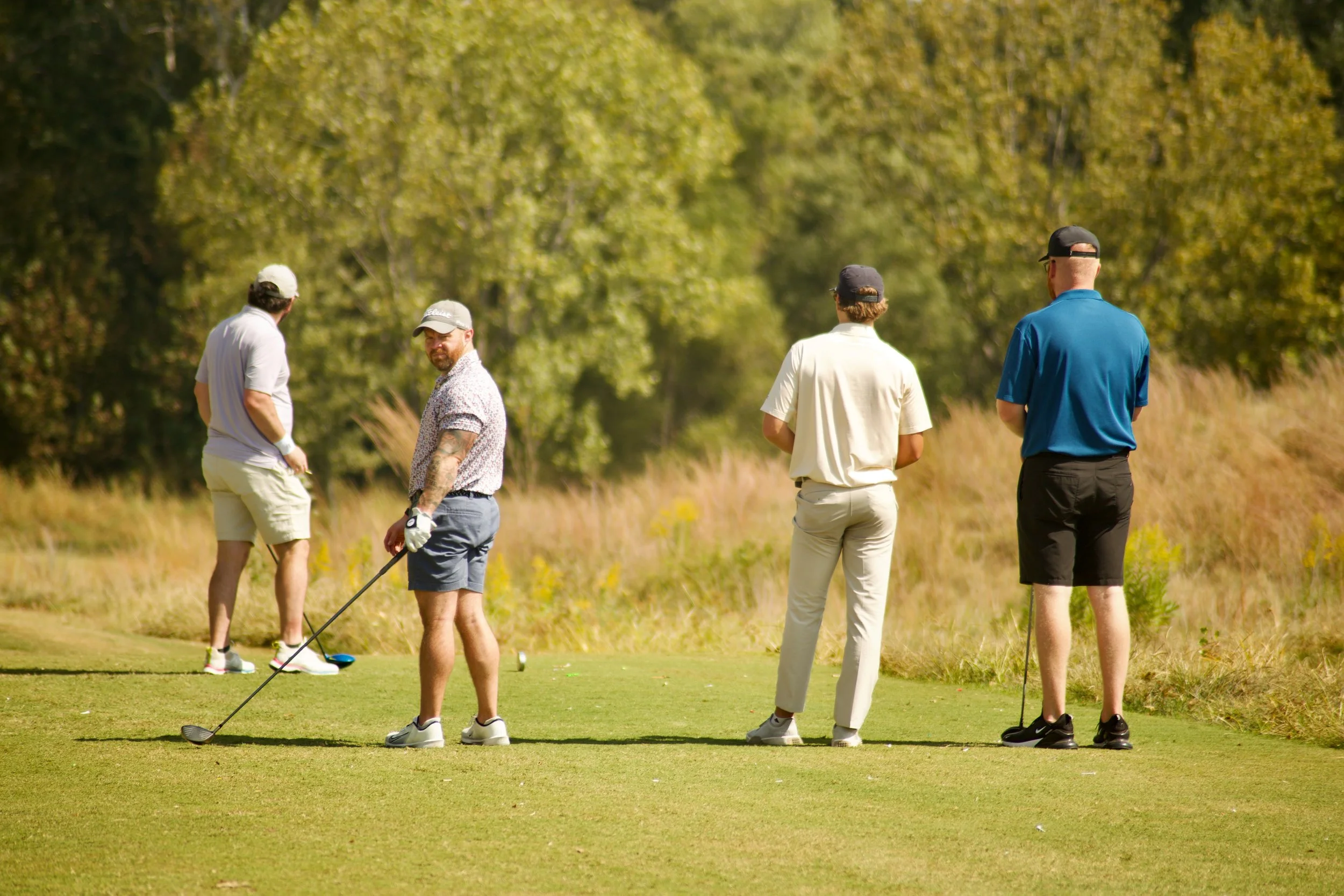 Four men playing golf on a sunny day, with one preparing to take a shot and the others standing nearby on the golf course.