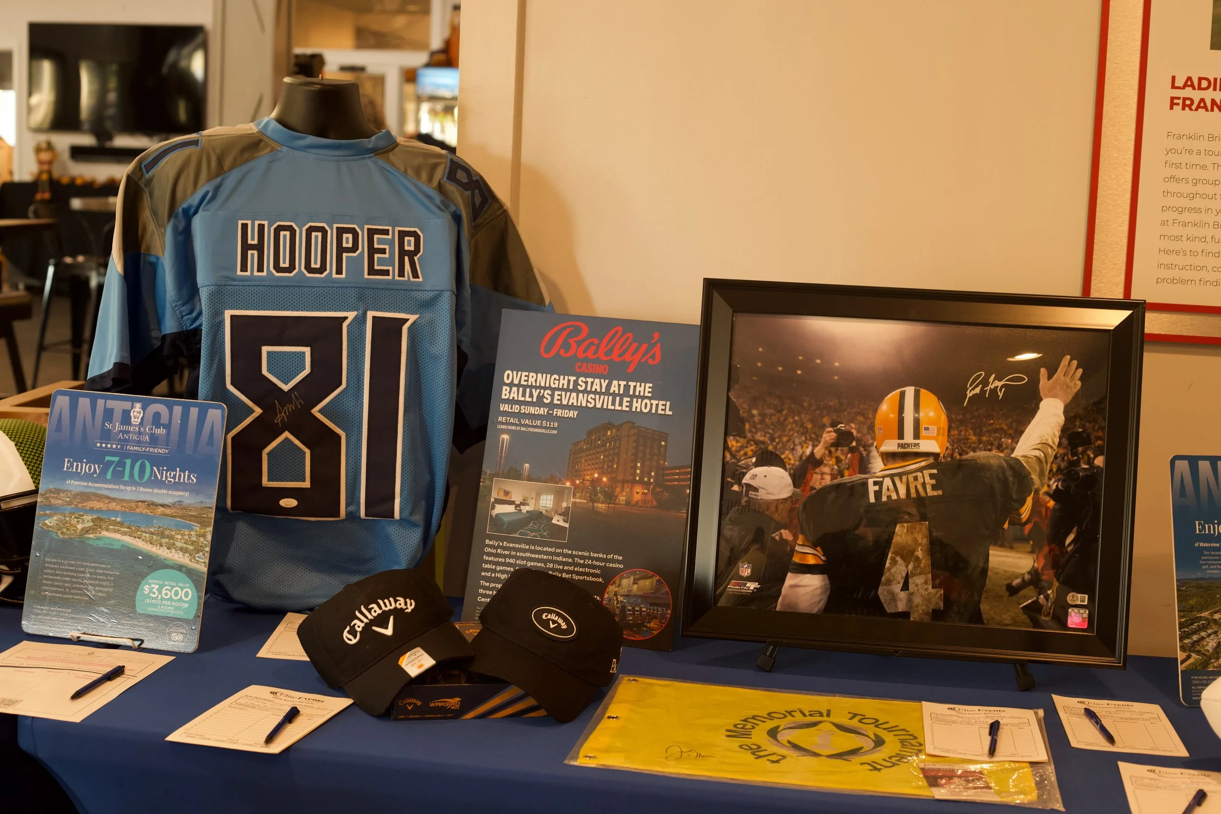 A display table with sporting memorabilia including a blue football jersey with the name 'HOOPER' and the number 81, framed sports photographs, promotional flyers for Bally's casino and Monte Carlo tour, baseball caps with Callaway branding, and info