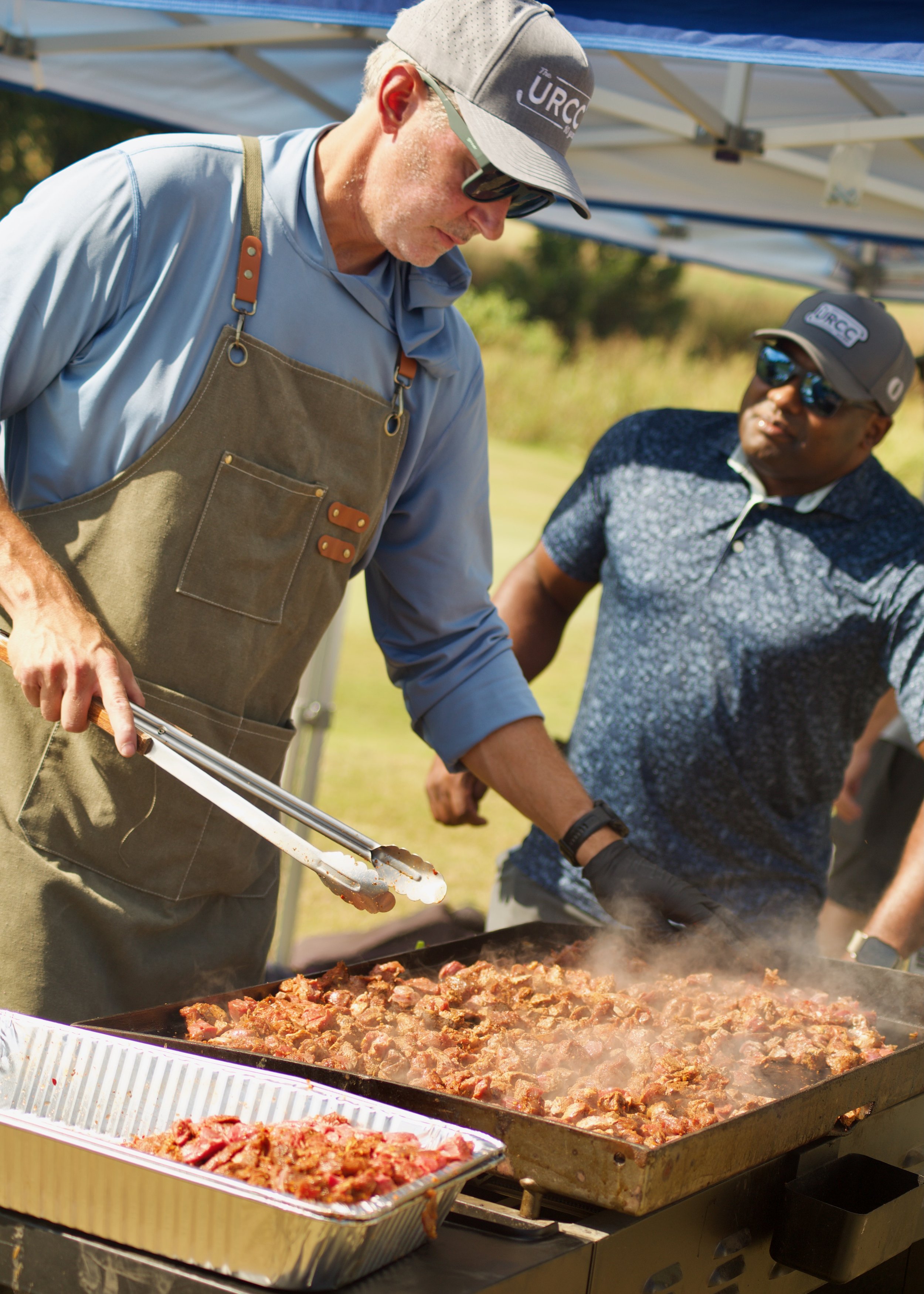 Two men cooking at an outdoor barbecue, one in a beige apron and cap, the other in a patterned shirt and sunglasses, roasting meat on a large grill under a canopy.