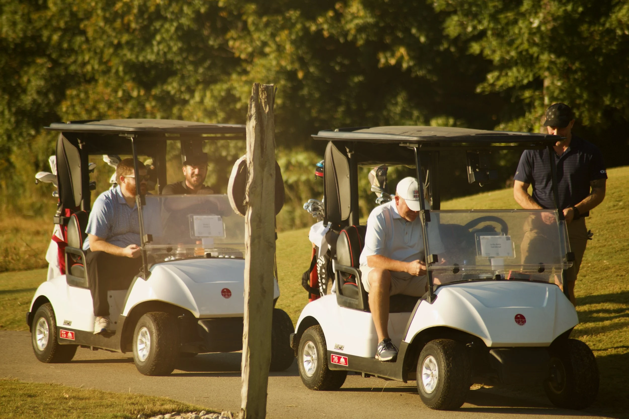 Several people riding in two golf carts on a golf course, with a tree in the foreground and green trees in the background.