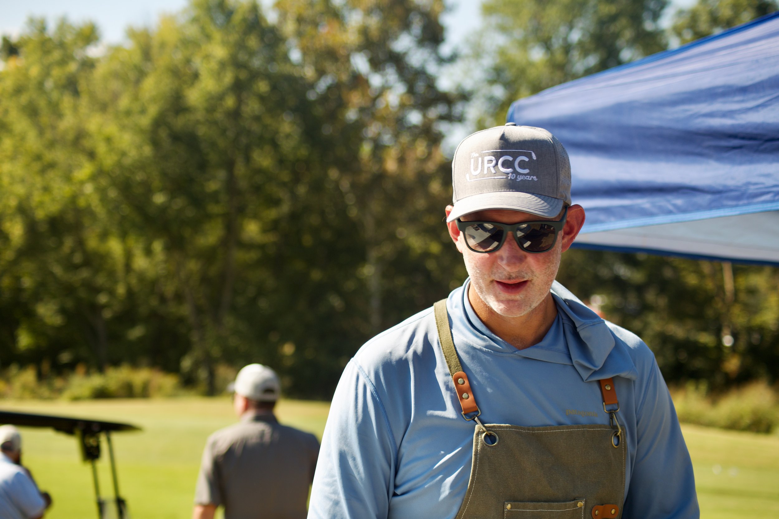 A man wearing sunglasses, a gray URCC cap, and a gray shirt with a brown apron outdoors during daytime, with trees and a blue canopy in the background.