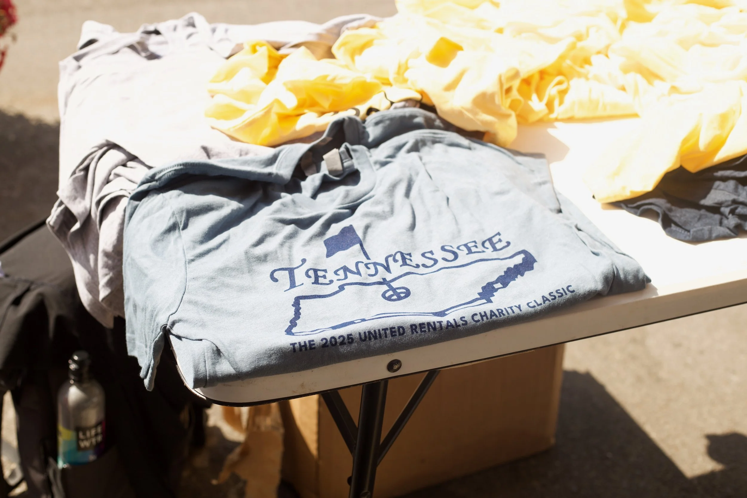 A table with folded shirts and yellow fabric, with a gray shirt displaying a Tennessee outline and text about the United Rentals Charity Classic event.