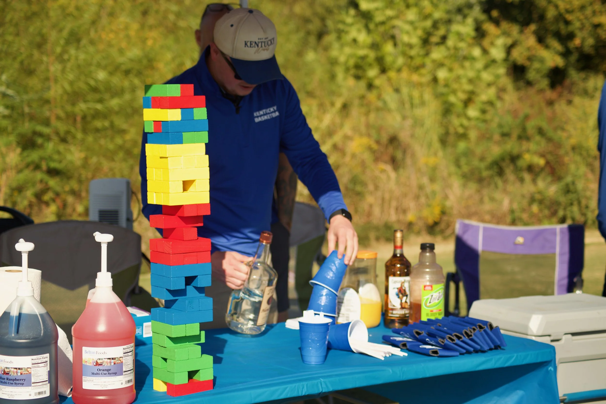 A person wearing a Kentucky basketball cap and a blue Kentucky basketball jacket stands outdoors at a table with large colorful Jenga blocks, bottles of syrup, paper cups, and other game supplies, with trees and green foliage in the background.