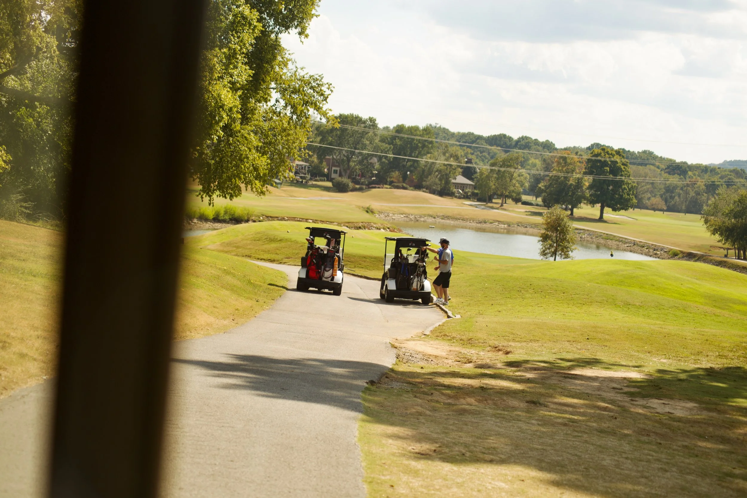 Golf course with two golf carts and a person standing between them, near a water hazard and surrounded by trees under a partly cloudy sky.