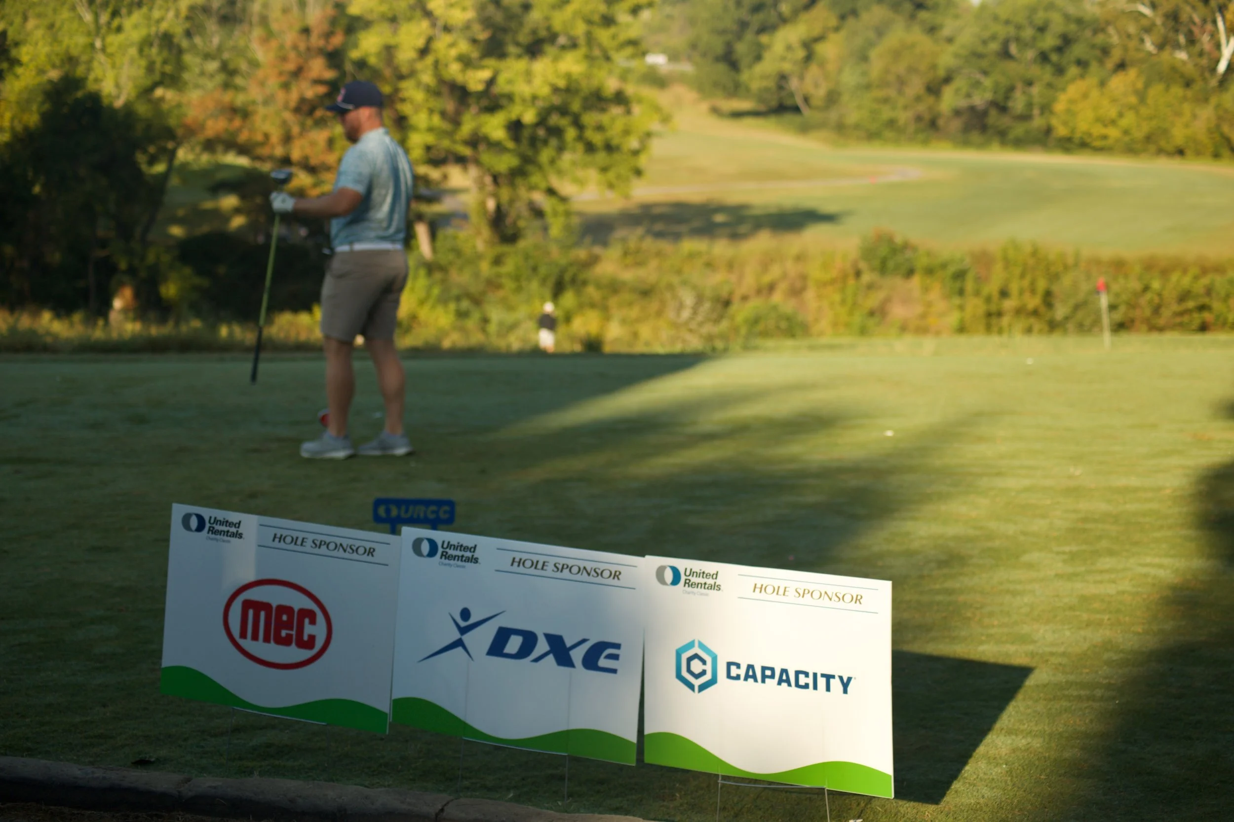 A man standing on a golf course putting green, wearing a cap, sunglasses, and golf gloves, surrounded by trees and a scenic landscape in the background. There are sponsor banners in the foreground.