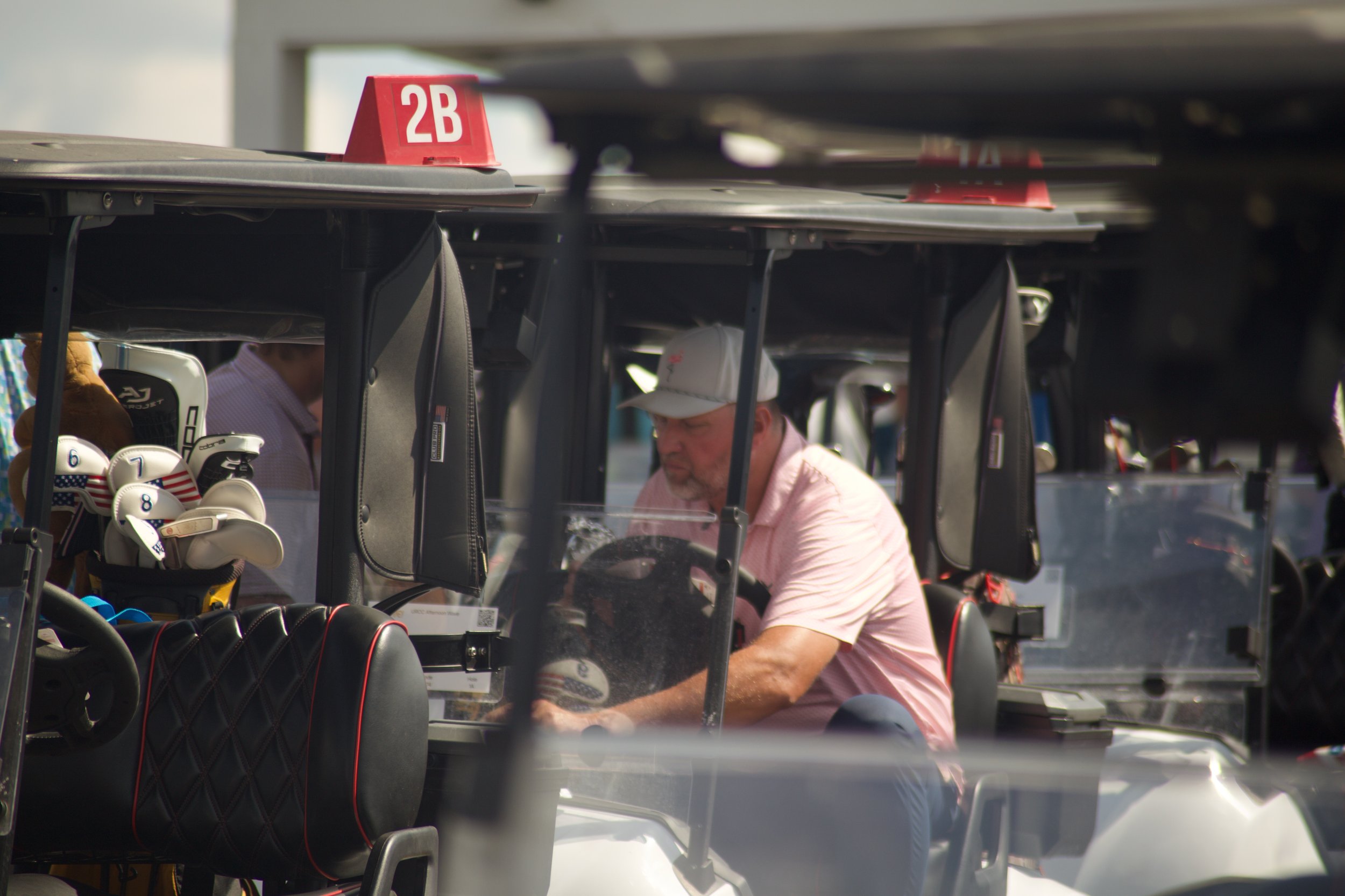Several golf carts parked in a row, with golf clubs and accessories visible, two men are inside one of the carts, one wearing a white cap and light pink shirt.