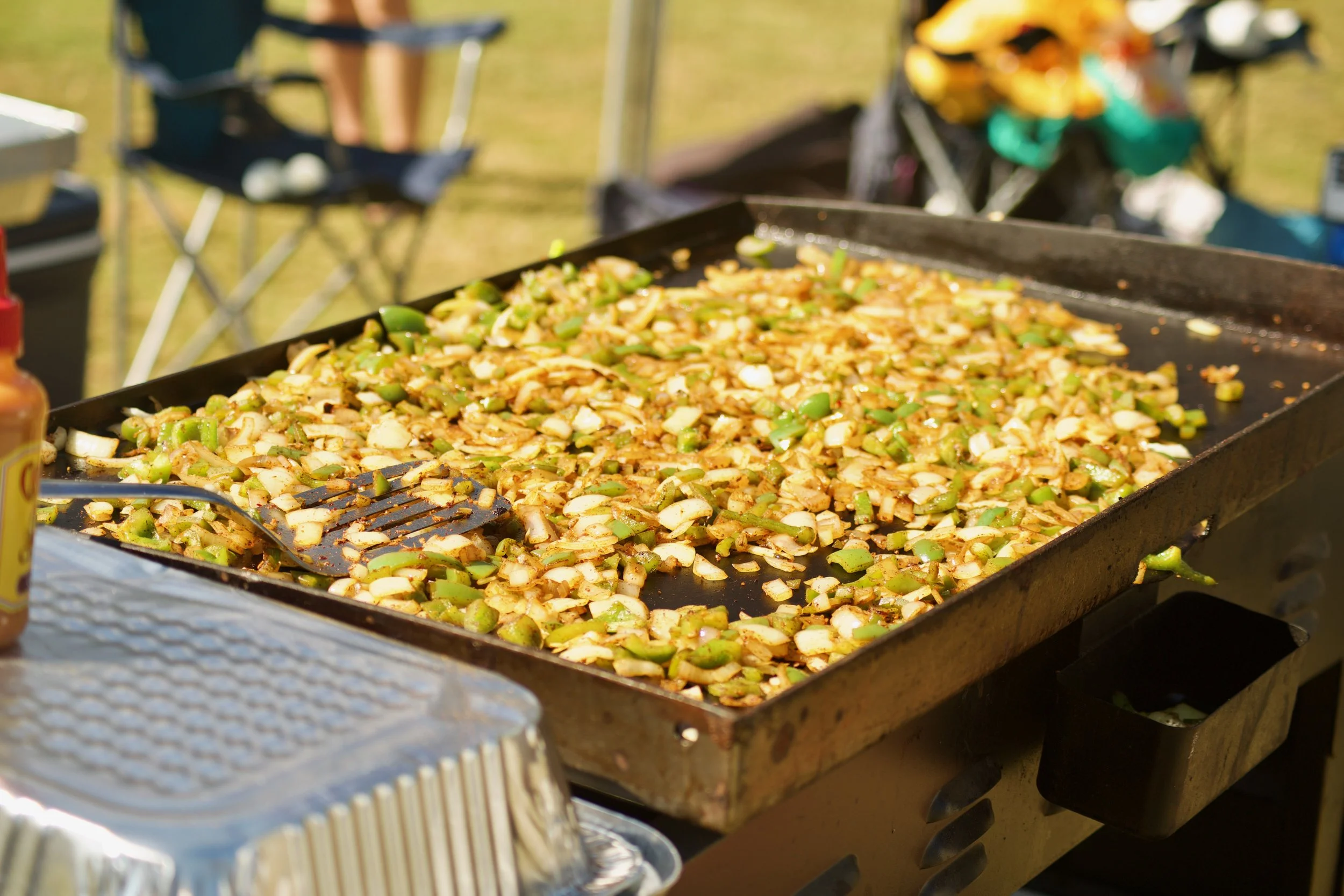 Grilled vegetables, including onions and green peppers, on a flat-top grill at an outdoor event.