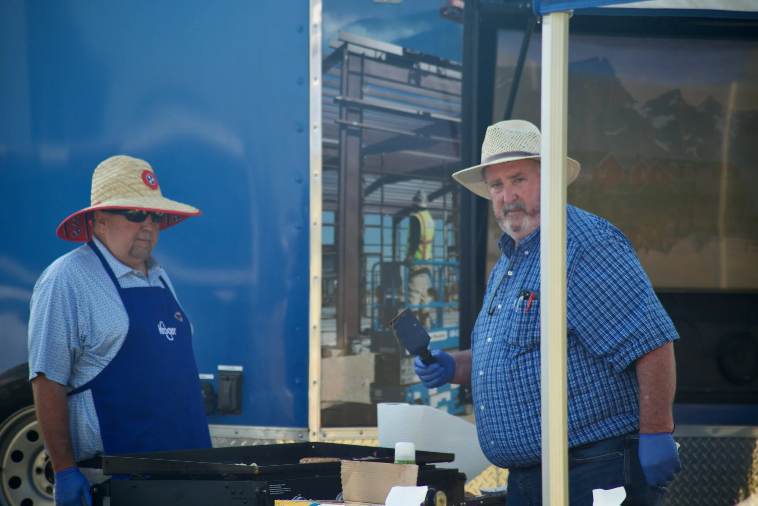 Two men with hats and blue shirts are standing outdoors near a food truck. One man is holding a tool and wearing gloves, while the other man wears sunglasses and a blue apron. They appear to be preparing or serving food.