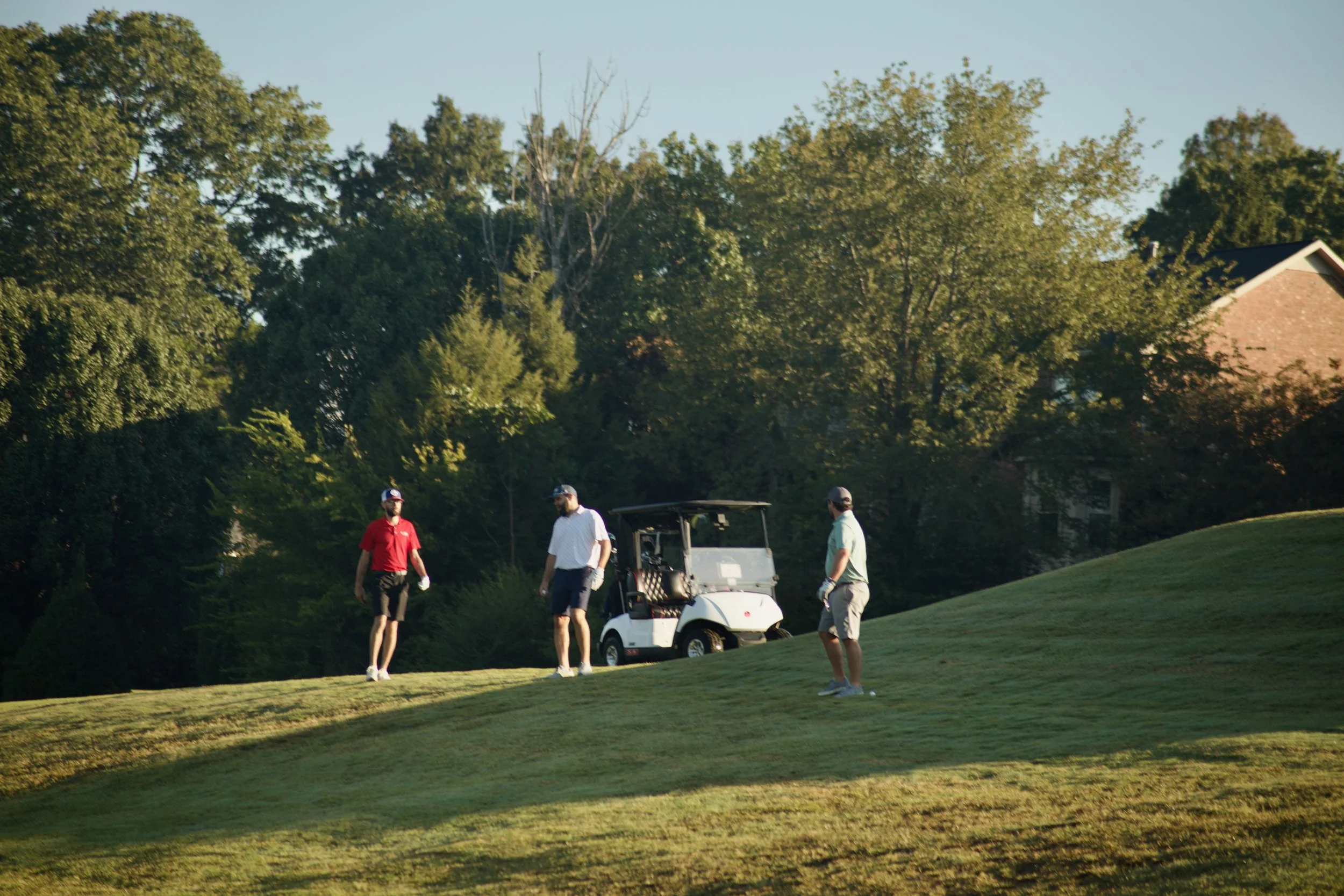 Four men are standing on a golf course near a golf cart, with trees and a house in the background. The men are dressed in golf attire, and the scene is lit by sunlight.