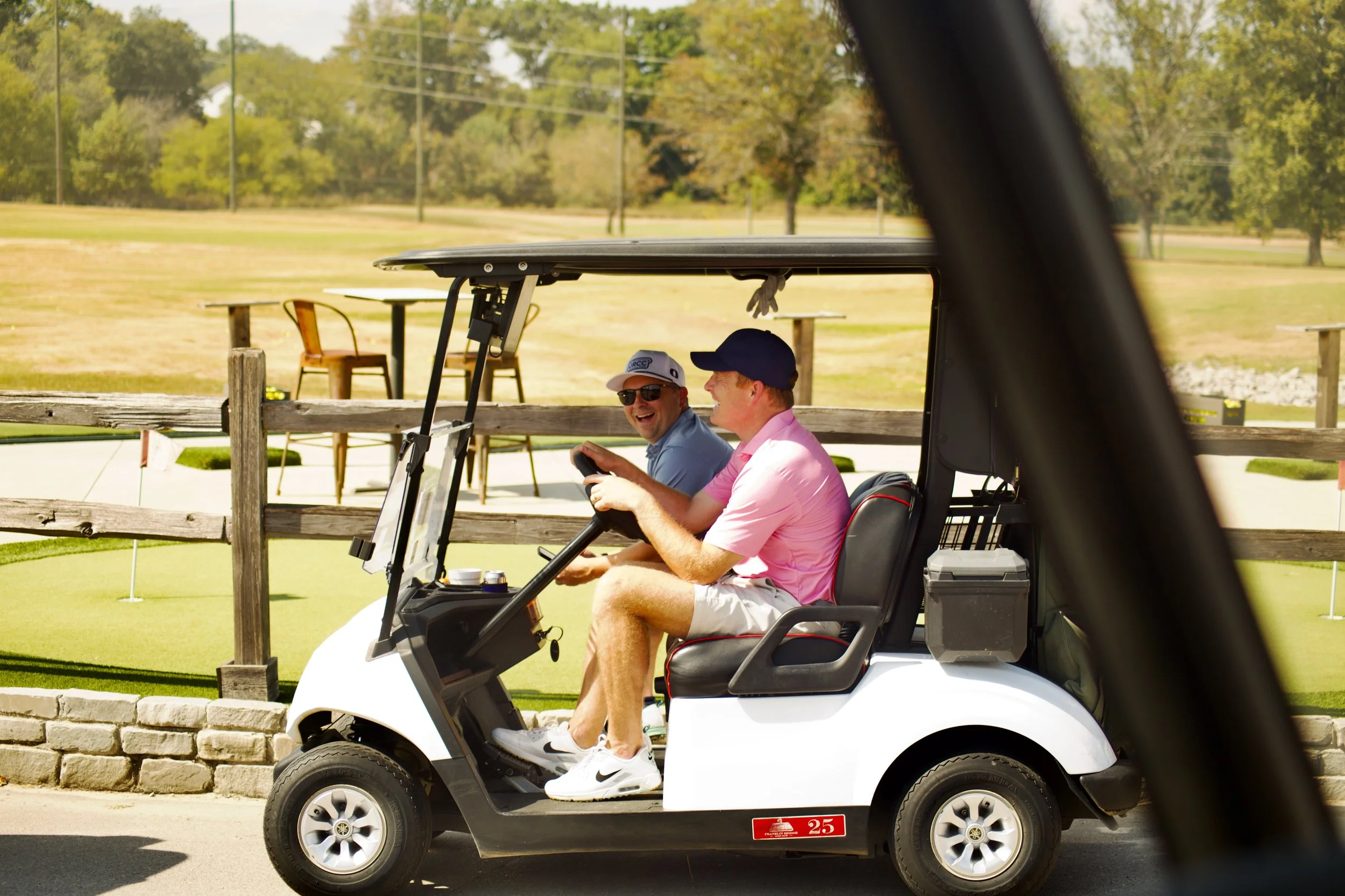Two men sitting in a golf cart on a golf course, laughing and talking, wearing casual golf attire and hats. The background shows trees and a grassy area.