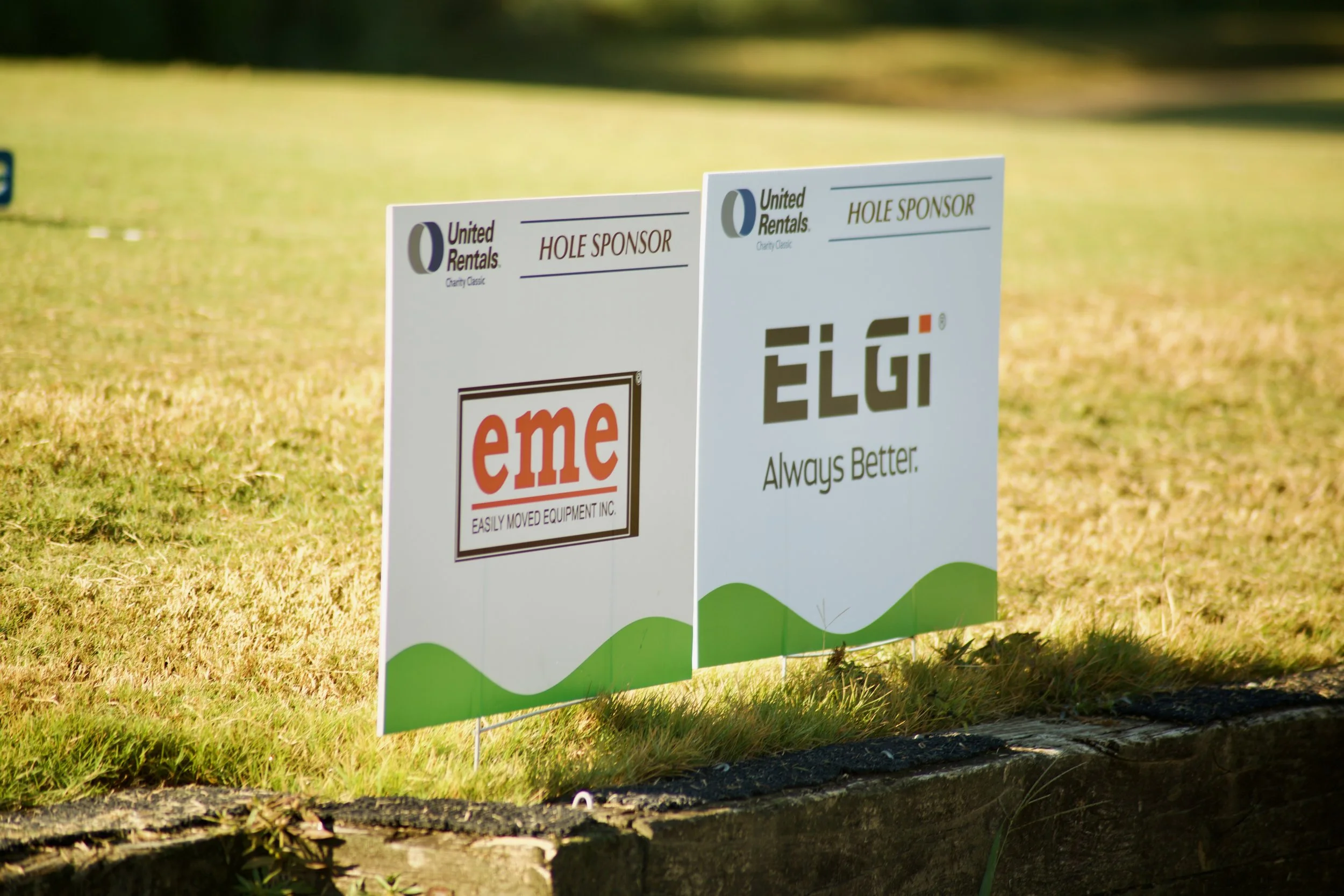 Signs on a grassy area for a golf tournament, sponsored by United Rentals, with logos and slogans for hole sponsorship and equipment rental companies.