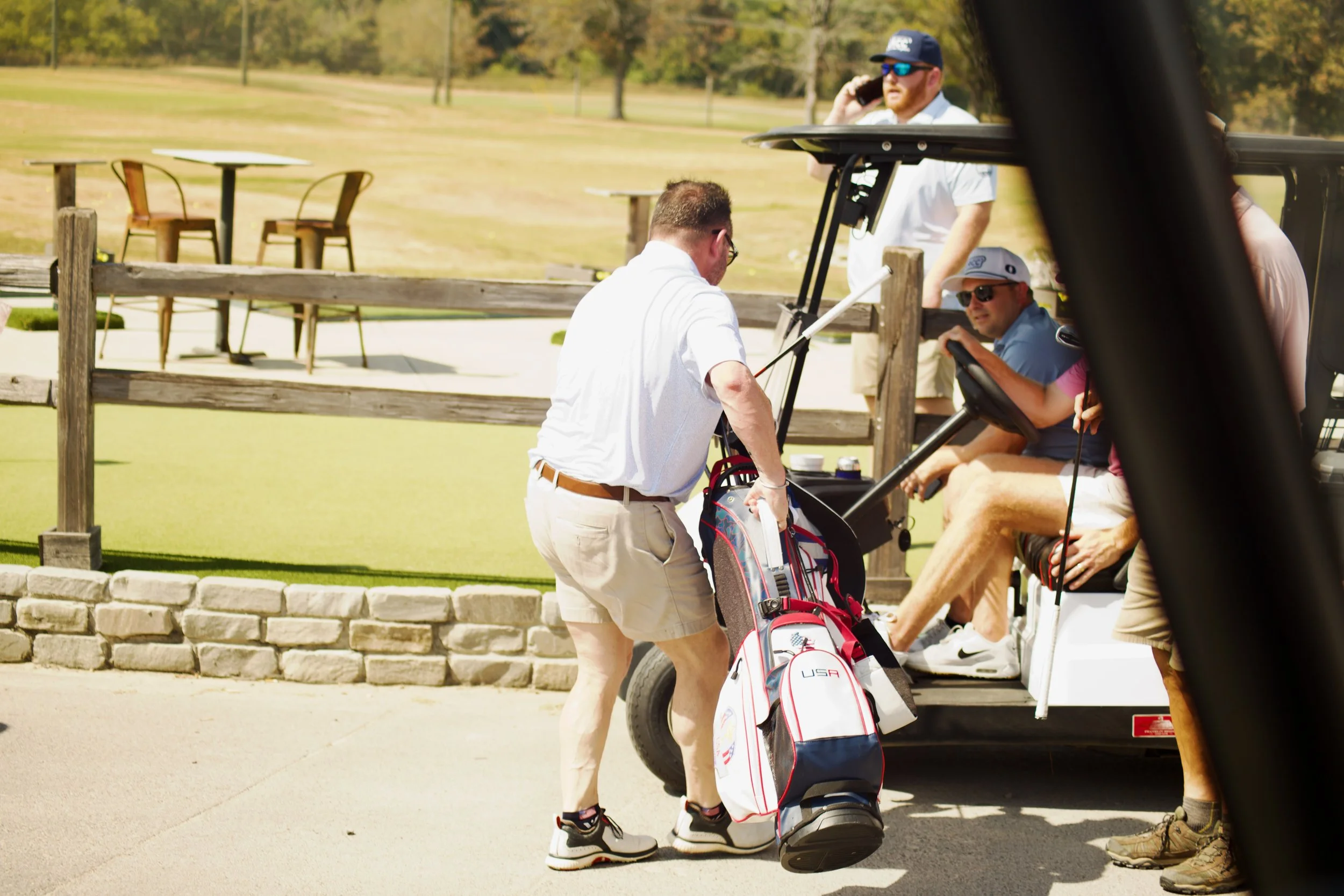 Men on a golf course preparing to play golf, with golf bags and a golf cart, in a park-like setting with trees in the background.