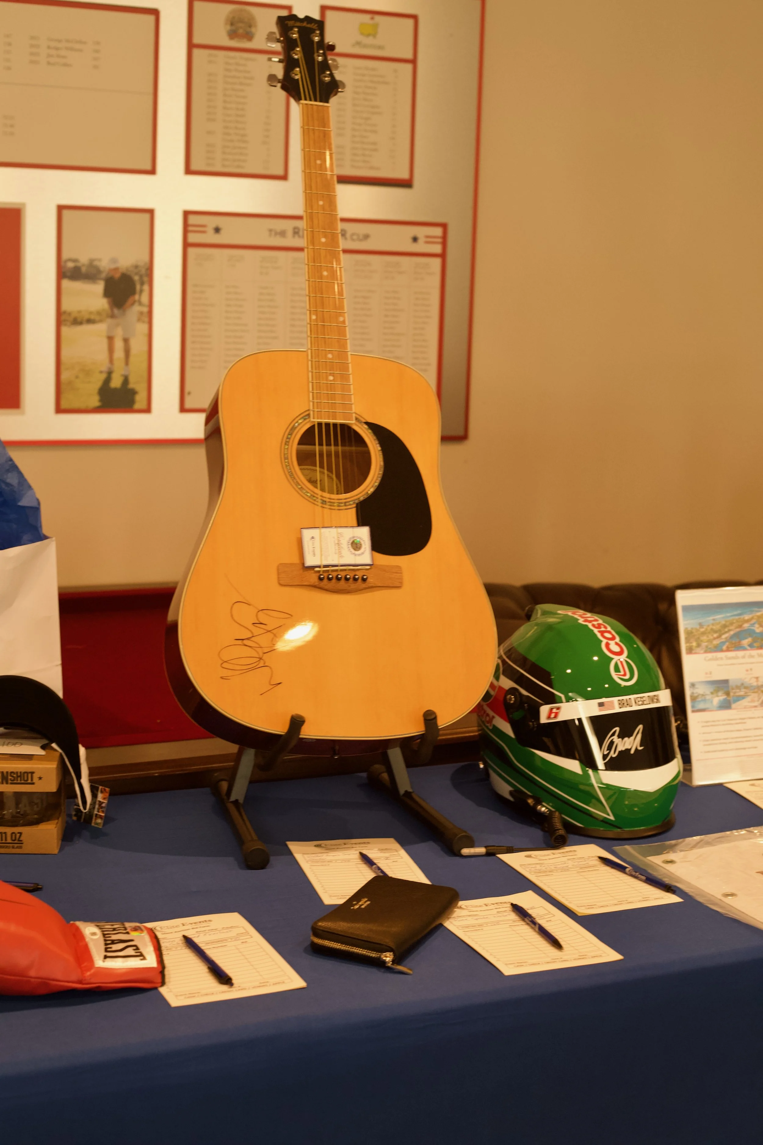 An acoustic guitar on a stand, a racing helmet, and informational materials on a table. In the background, posters and a man in shorts are visible.