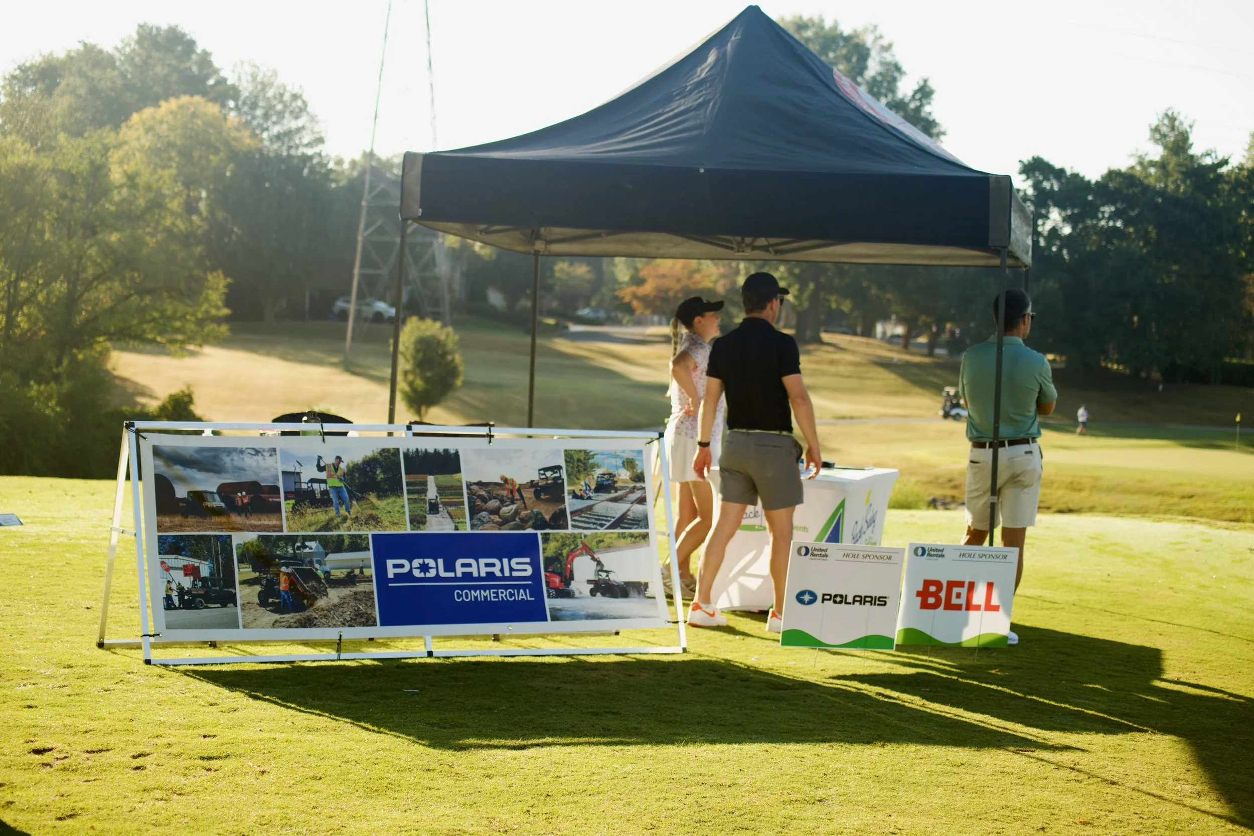 An outdoor event on a golf course with a tent, people, and promotional banners for Polaris, Bell, and United Rentals, with trees and a transmission tower in the background.