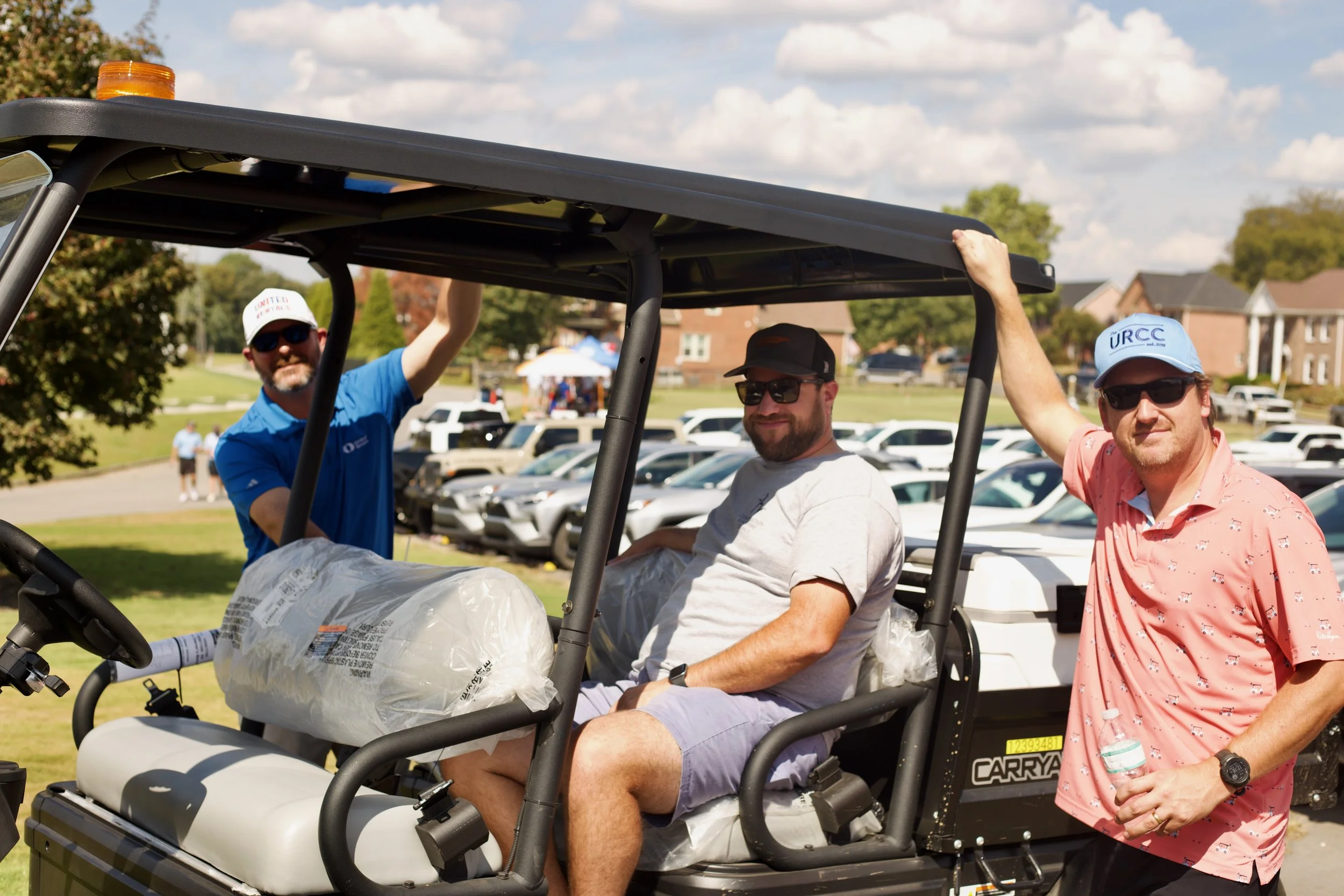 Three men sitting in a golf cart, with a woman standing beside them, all outdoors on a sunny day with houses and cars in the background. The man on the left is wearing sunglasses, a cap, and a blue shirt. The man in the middle is wearing sunglasses, 