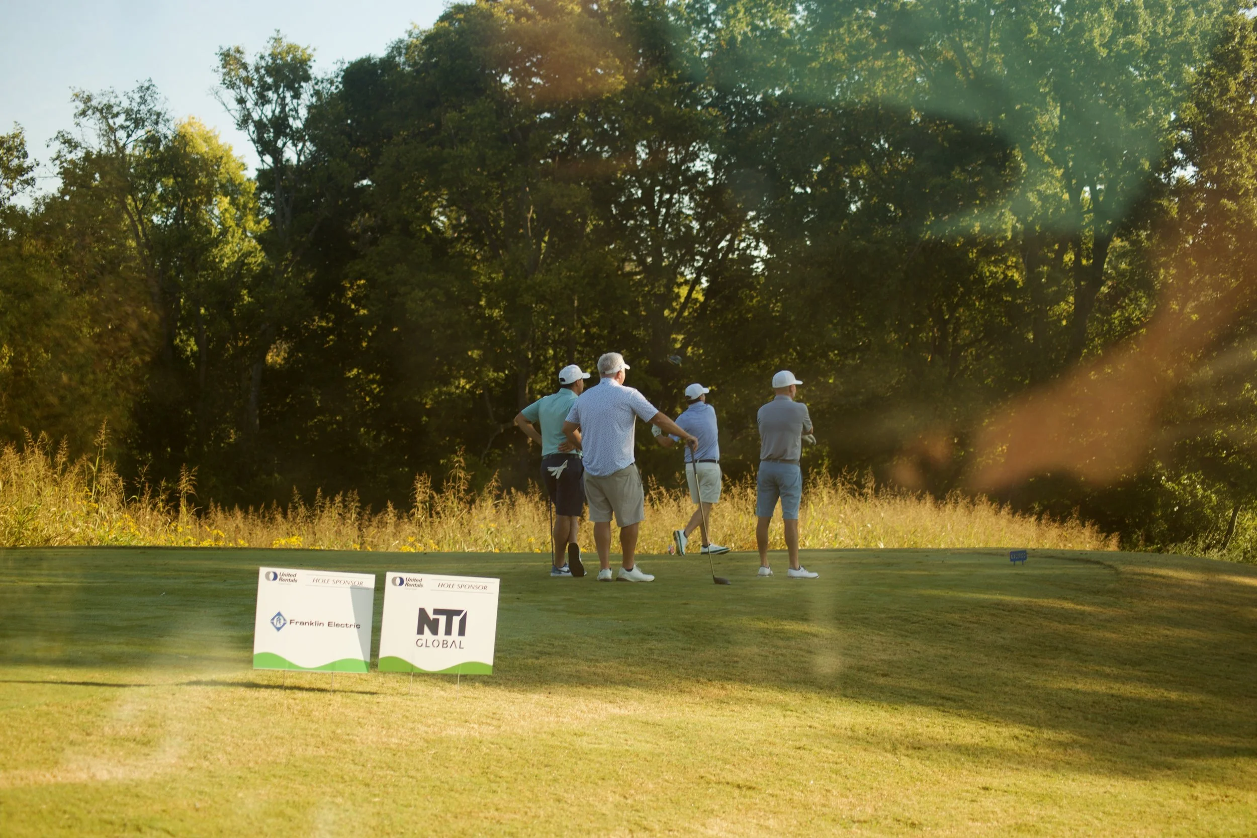 A group of five men playing golf on a course surrounded by trees in the late afternoon or early evening. Two signs with sponsor logos are visible in the foreground. The men are wearing golf attire and hats, and some are holding golf clubs.