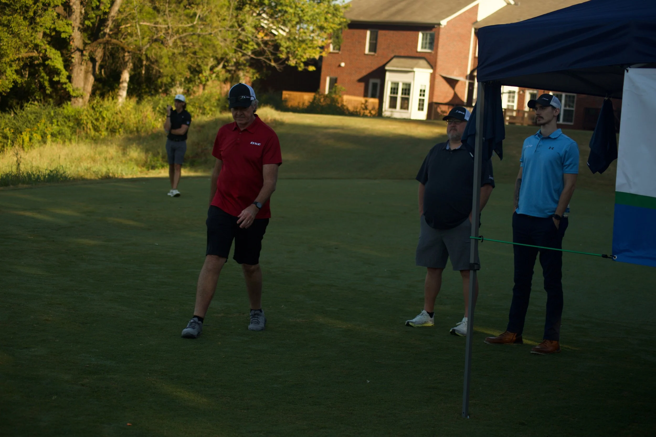 Four men on a golf course near a small tent, with a residential building in the background. One man in a red polo shirt walks in foreground, others standing nearby.