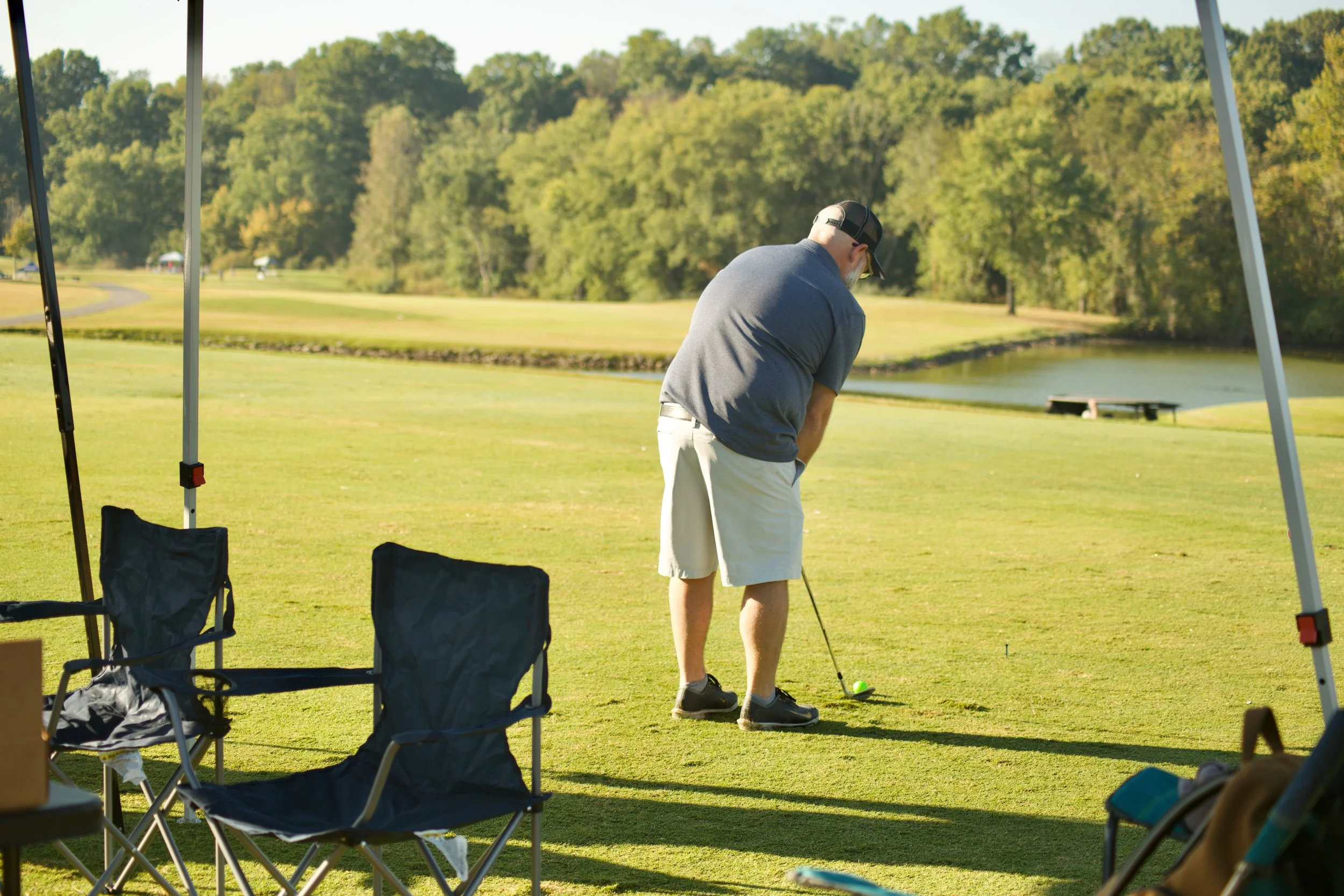 Man playing golf on a lush green golf course with trees in the background, viewed from under a canopy beside camping chairs.