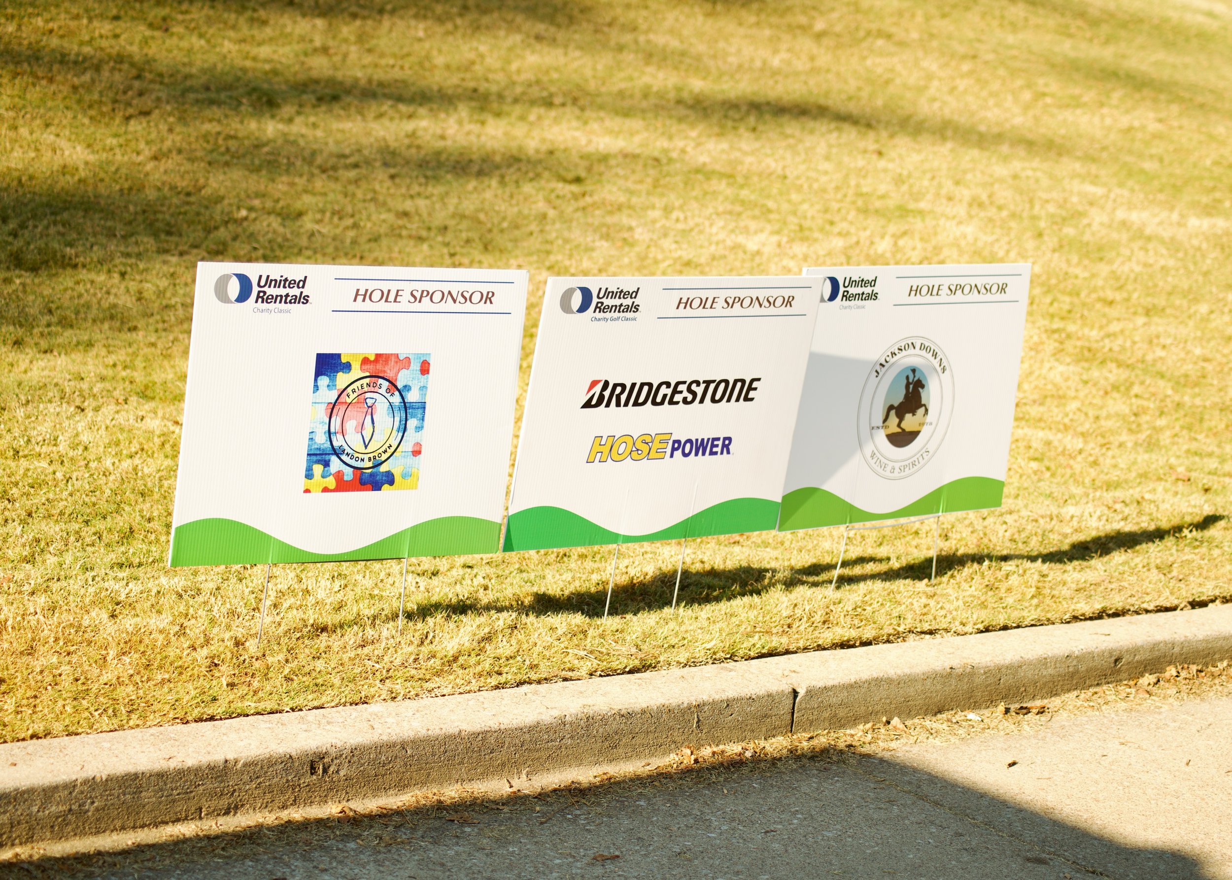 Three signs on a grassy area near a sidewalk, with logos and text indicating sponsorship for a charity golf event.
