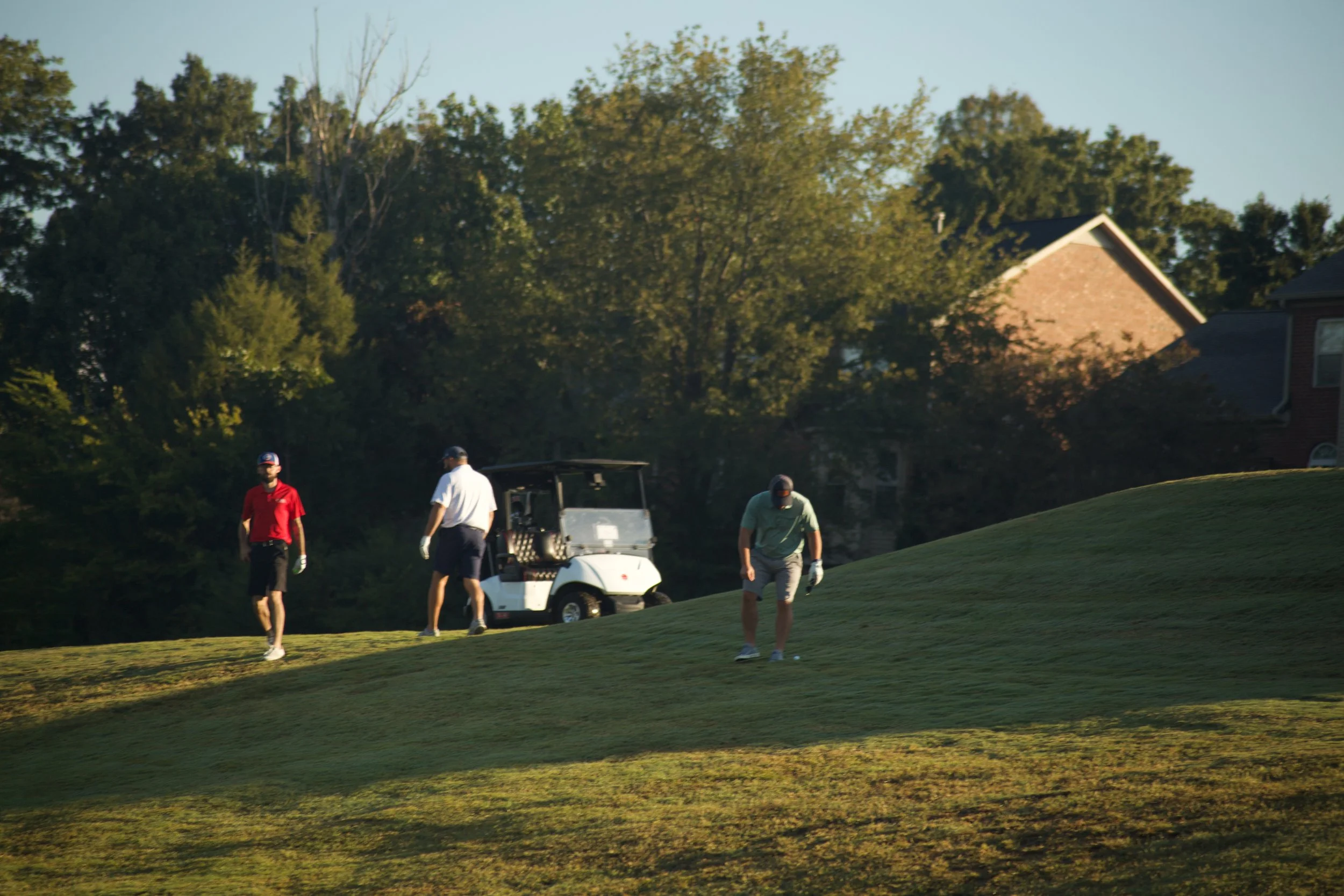 Several men playing golf on a lush green golf course, with a golf cart and trees in the background during late afternoon.