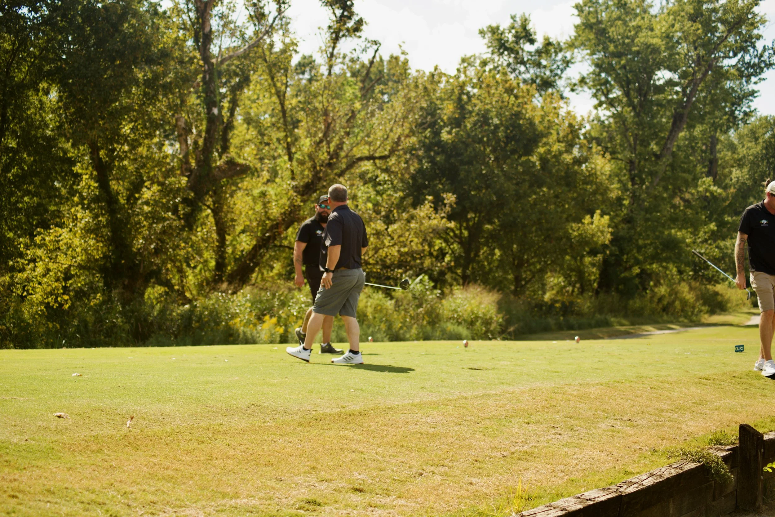 People playing golf on a lush green course surrounded by trees on a sunny day.
