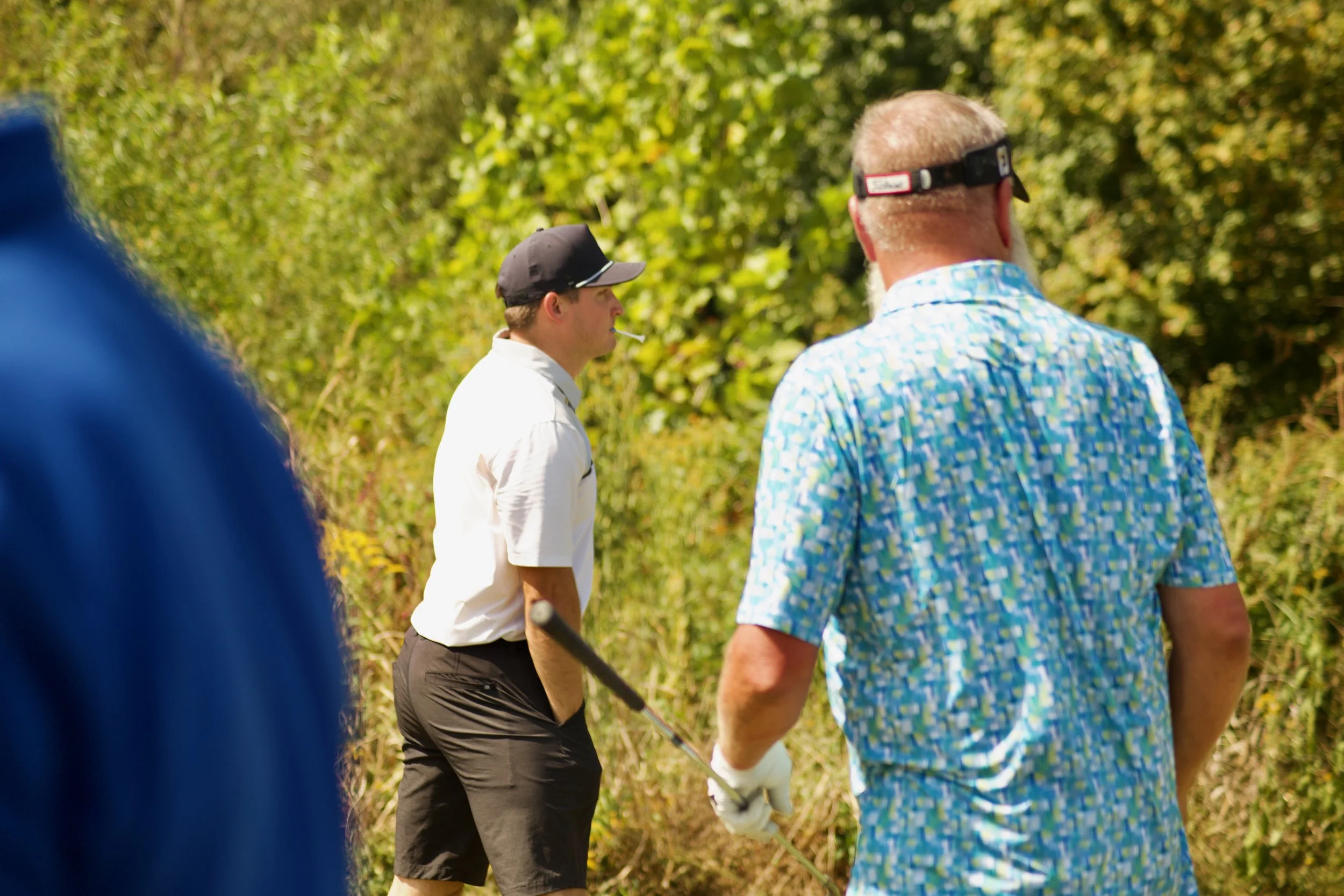 People are standing outdoors on a sunny day, with trees and green foliage in the background, engaging in conversation or socializing.