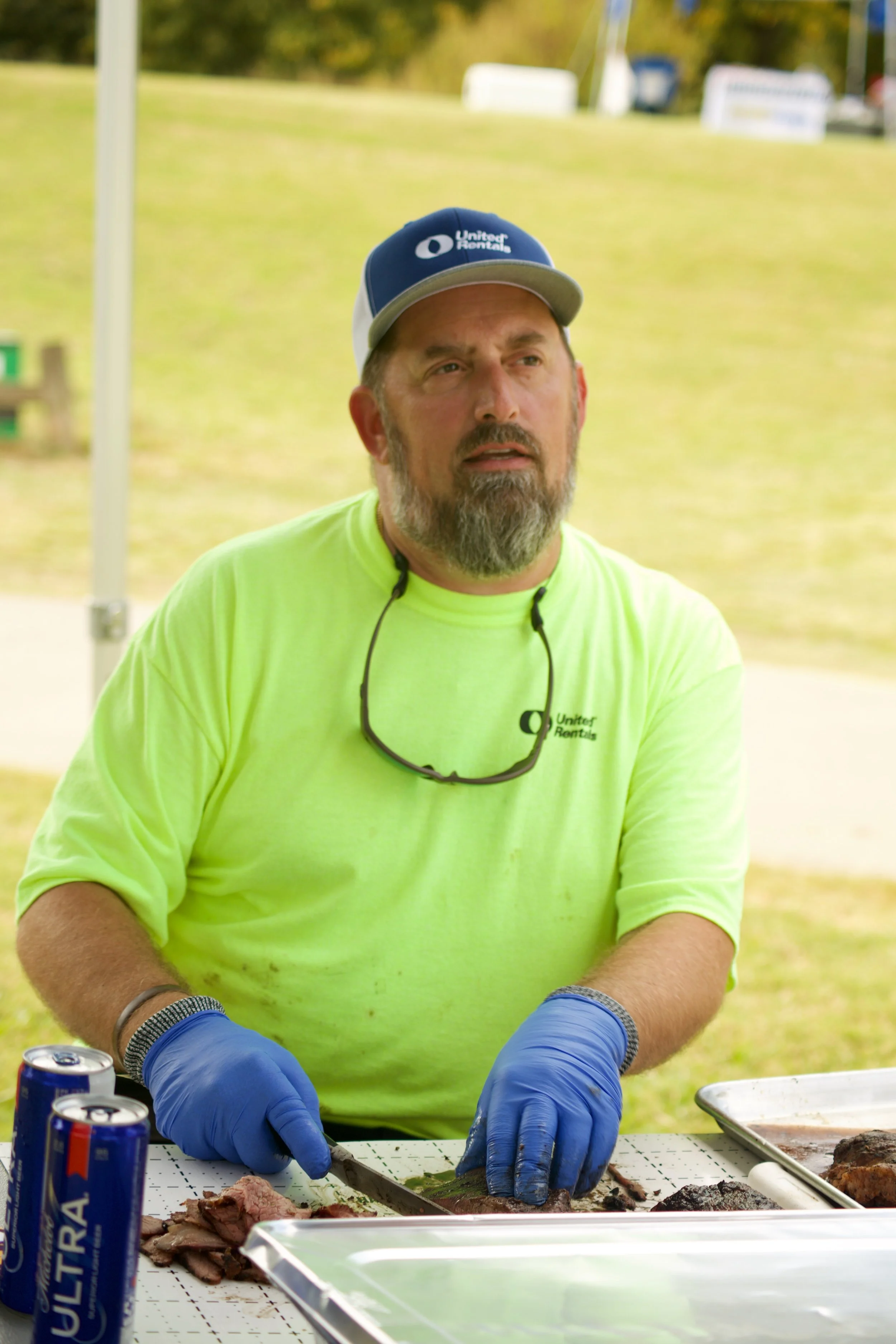 A man wearing a neon yellow shirt, blue gloves, and a United Rentals cap is slicing cooked meat at an outdoor barbecue event. There are soda cans and trays of food on the table.