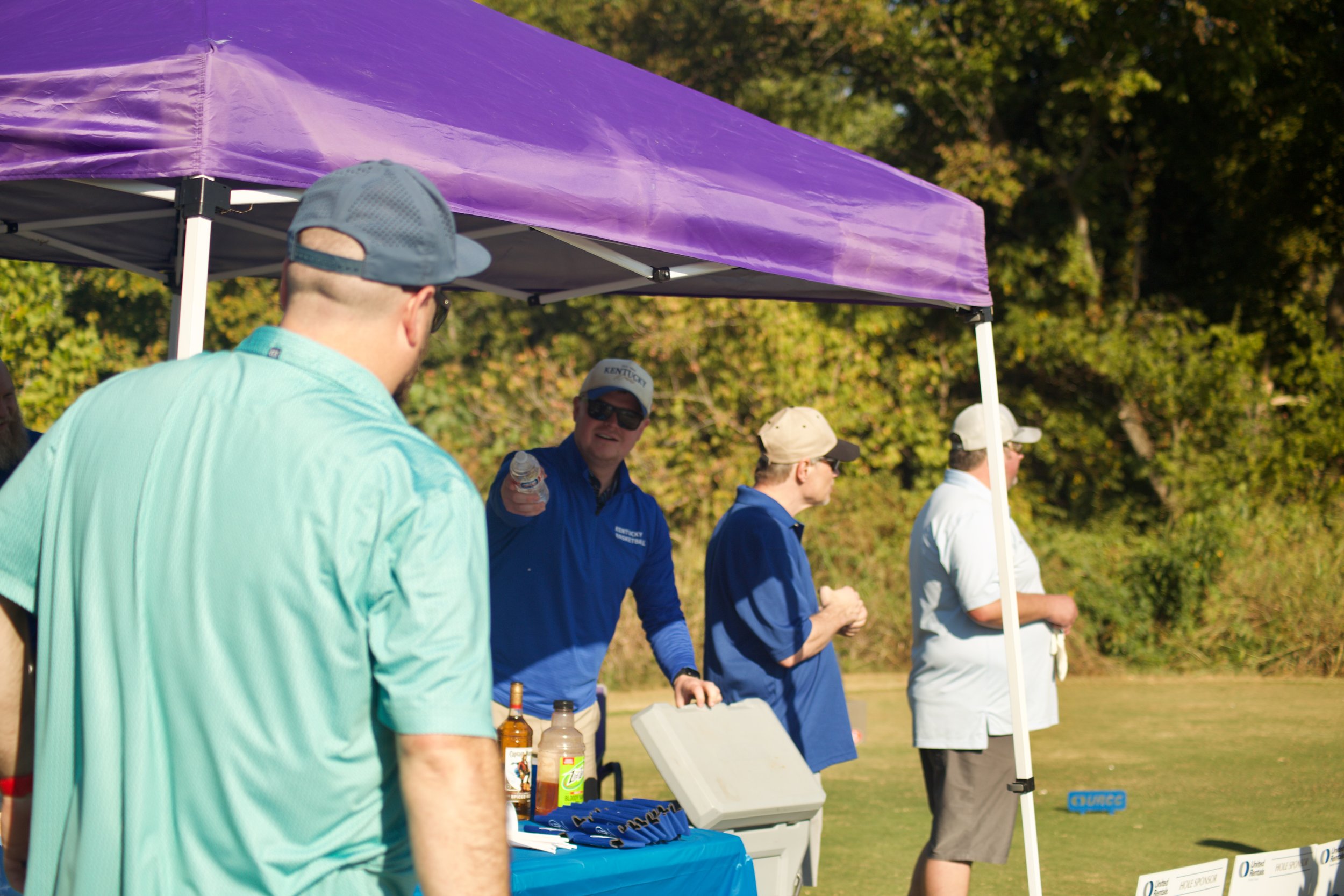 Group of people standing under a purple canopy outdoors with trees in the background, engaging in an event or gathering.