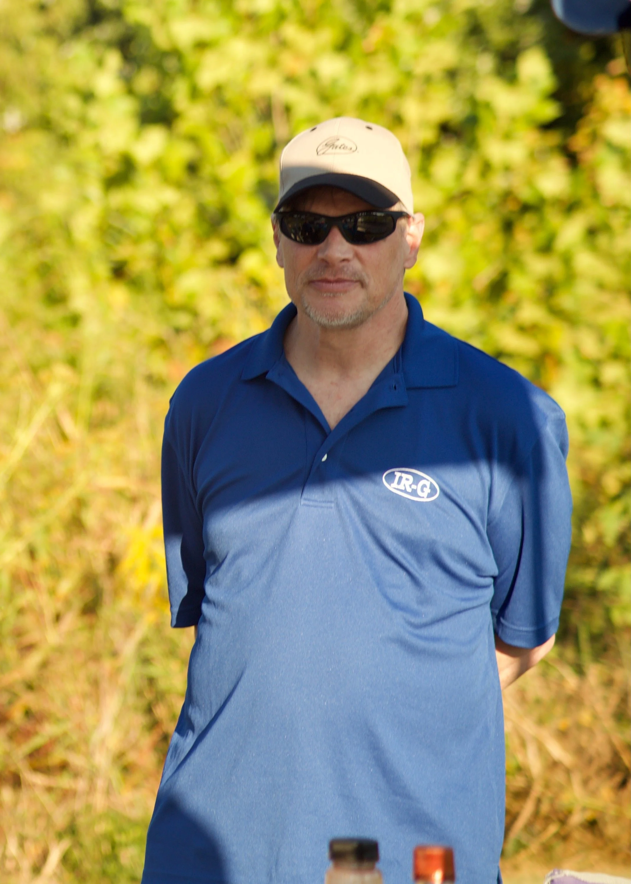 A man wearing dark sunglasses, a beige cap, and a blue polo shirt standing outdoors with a blurred background of green foliage.