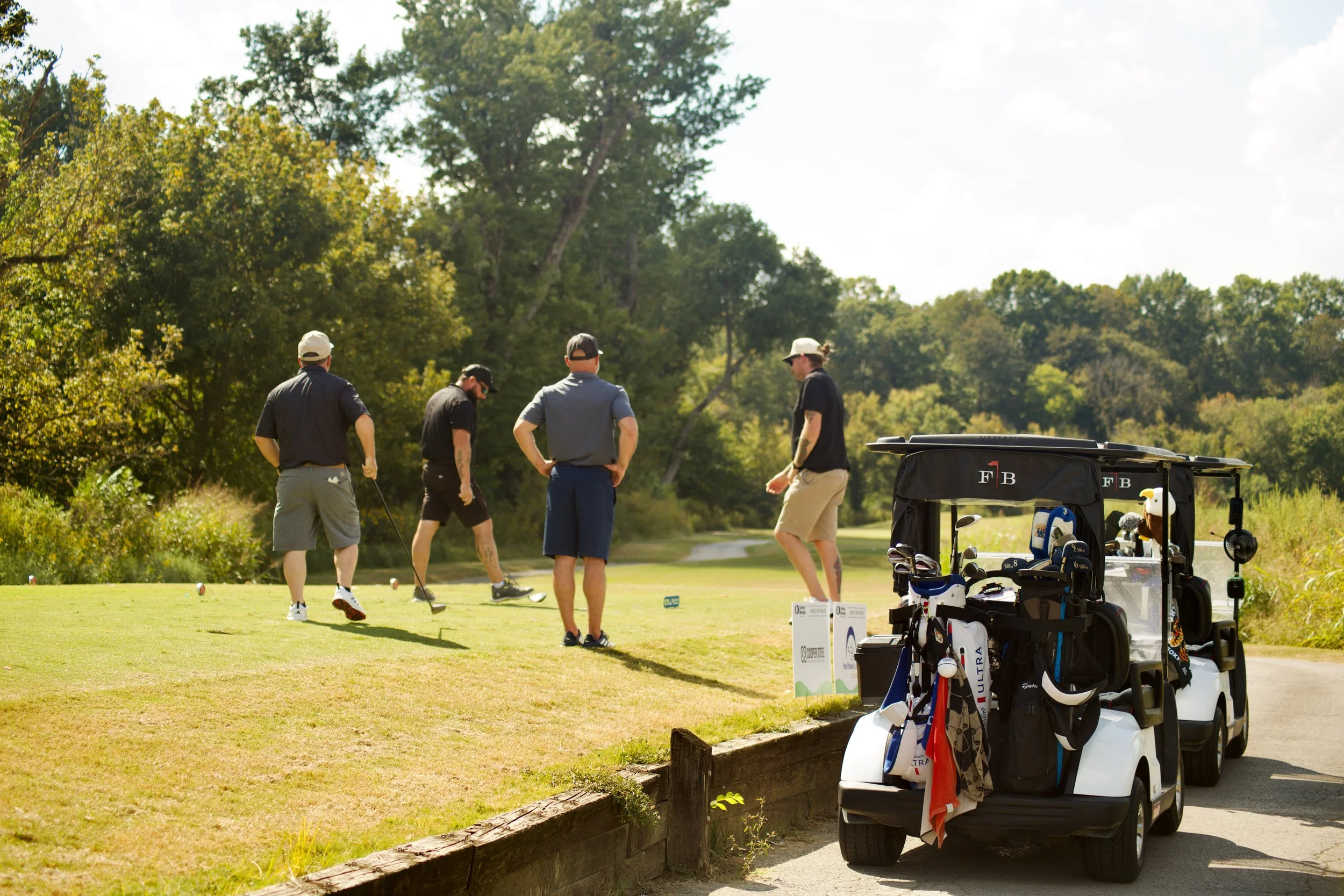 A group of people playing golf on a sunny day with trees in the background and a golf cart loaded with golf clubs in the foreground.