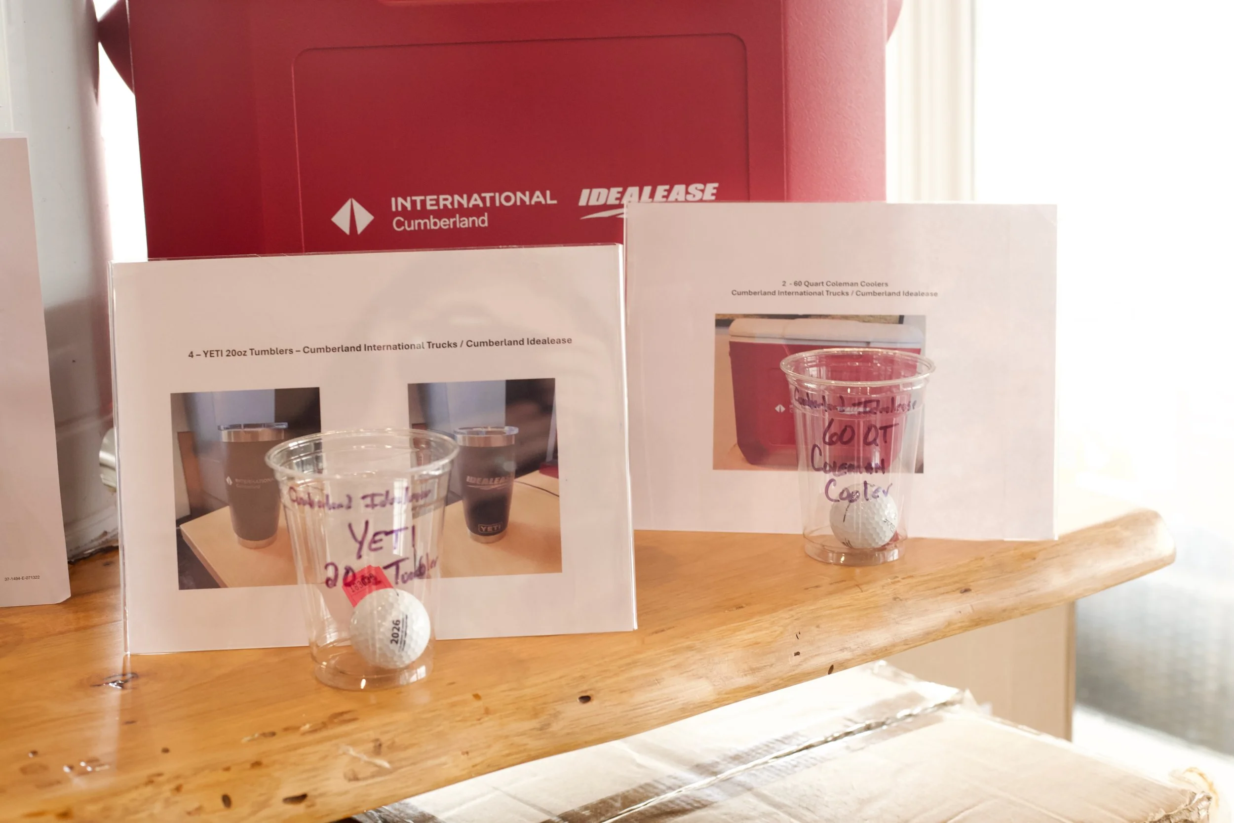 Display of two printed sheets on a wooden surface, each showing an image of a Coleman cooler and a cup with a golf ball on top. The cups have handwritten labels indicating they contain iced coffee and are priced at $6.00.