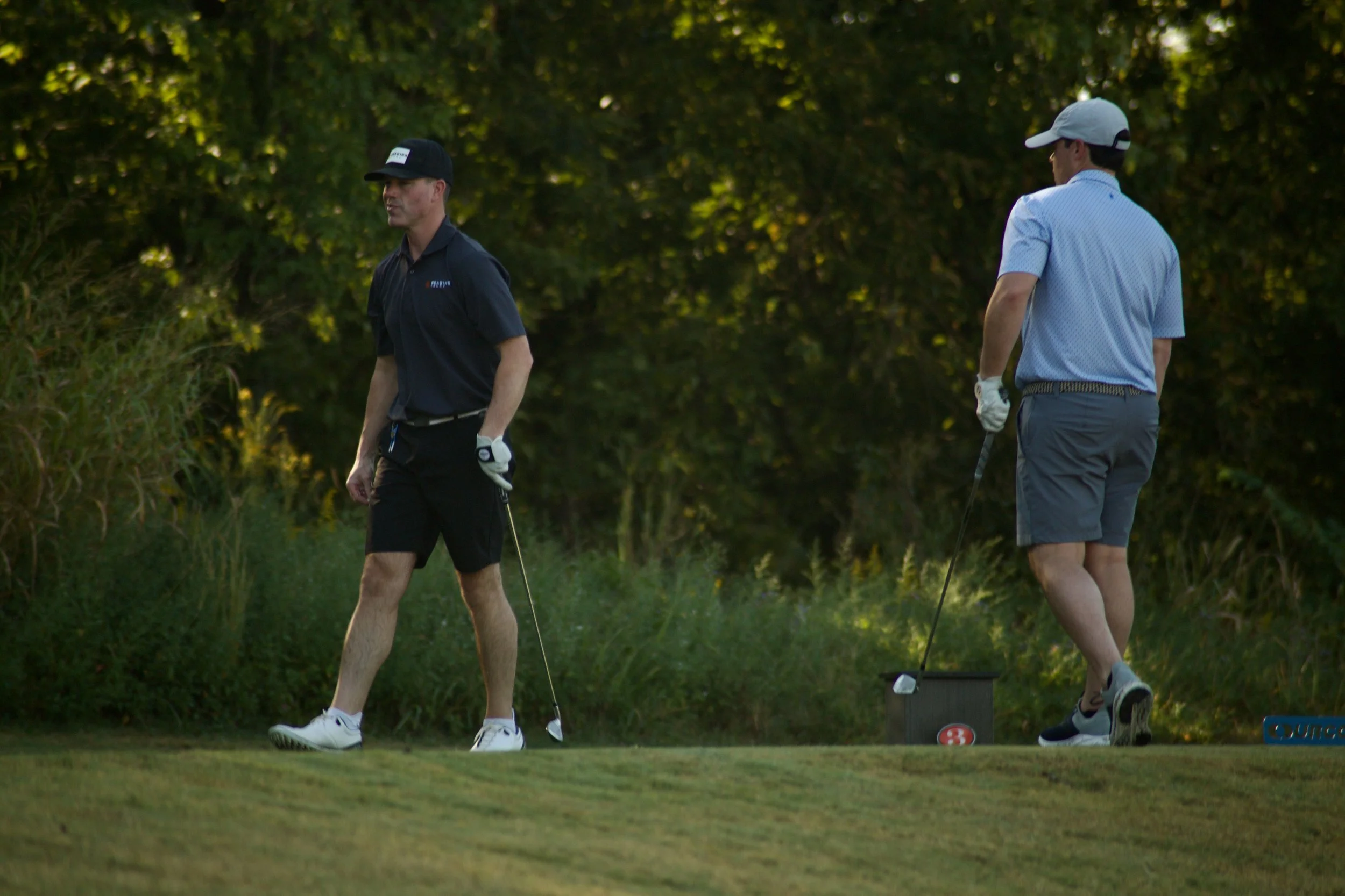 Two men playing golf on a lush green course during daytime with trees in the background. One man is wearing a dark shirt and black hat, the other is dressed in a light blue shirt and white cap.