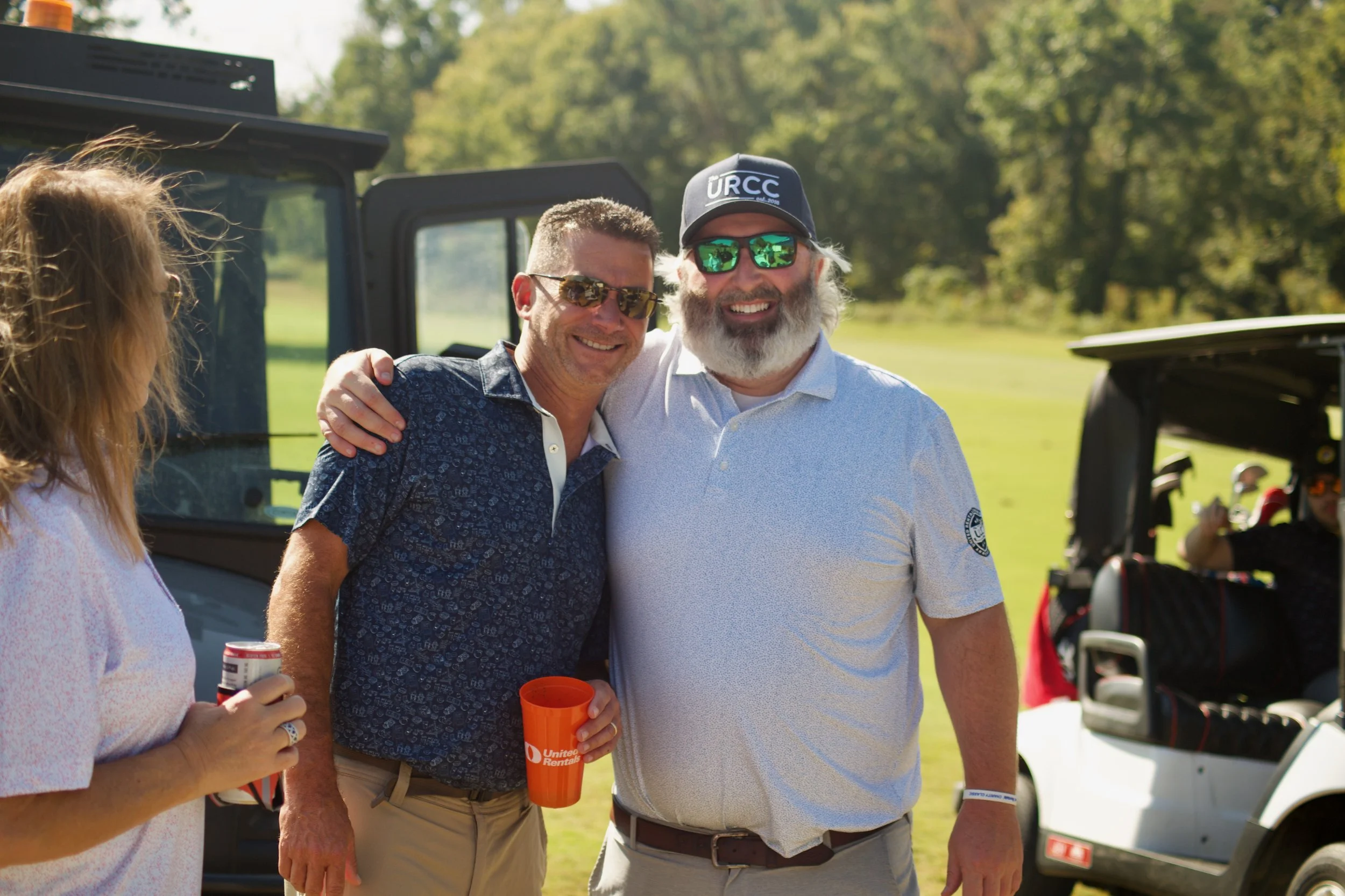 Group of people outdoors on a sunny day, smiling and posing for a photo. Two men are in the center, one with a beard and wearing sunglasses, the other with short hair and glasses. A woman is partially visible on the left holding a beverage. There is 