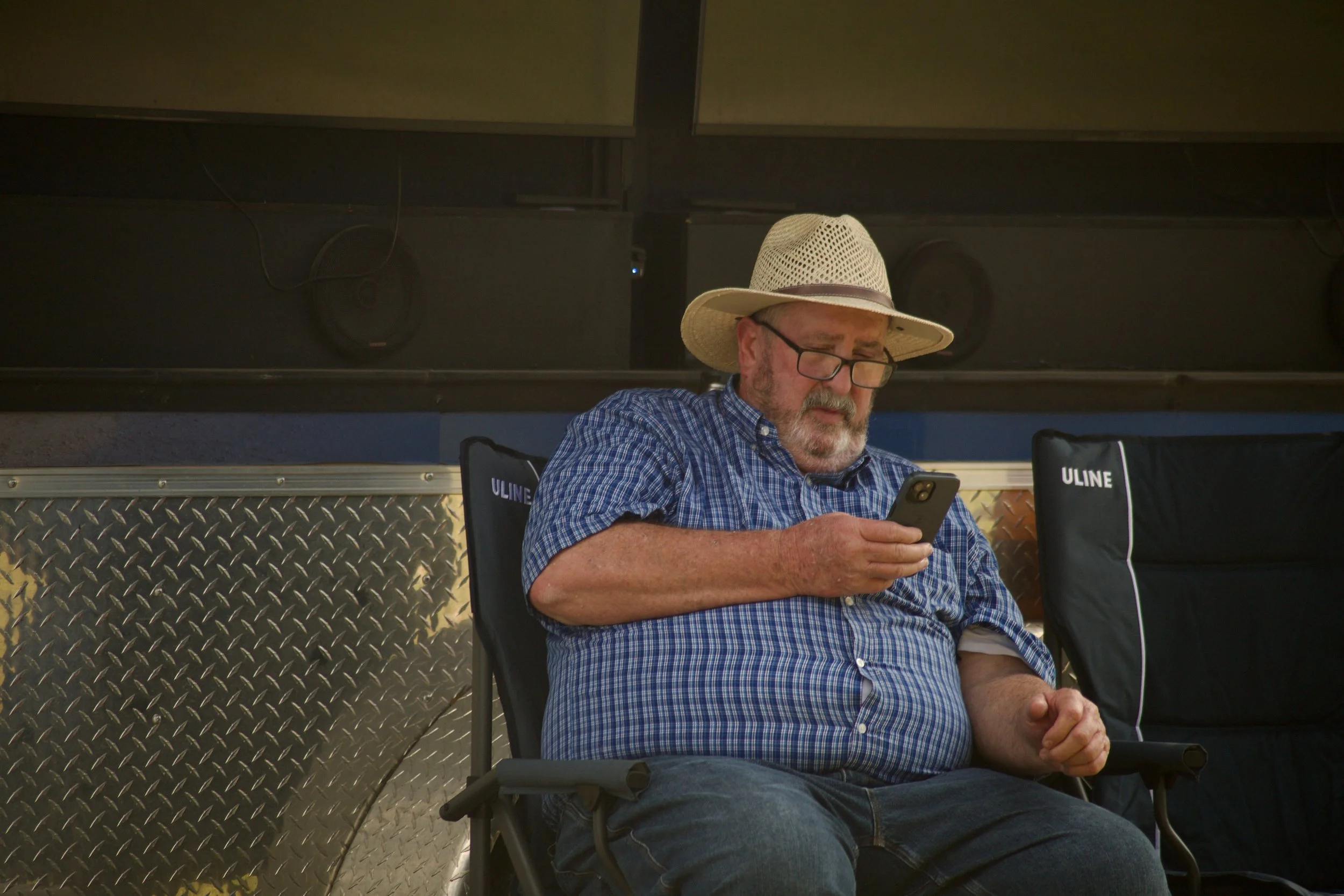 A man wearing a straw hat, glasses, a blue checkered shirt, and jeans sitting in a camping chair, looking at his phone, with a black and white cushion nearby, in a setting with a black ceiling, speakers, and a metallic textured wall.