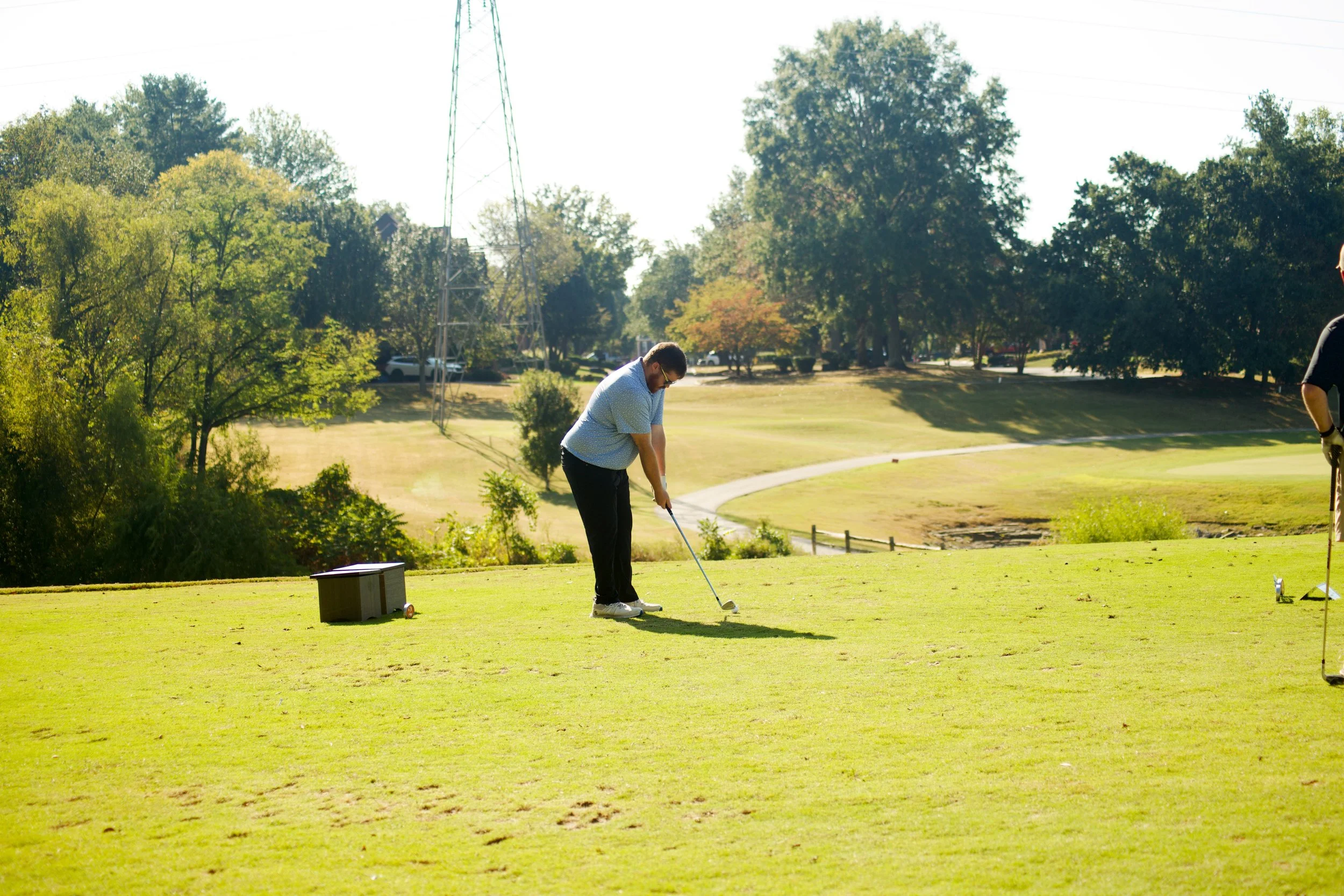 A person preparing to hit a golf ball on a golf course during the day. There are trees and a power line in the background.