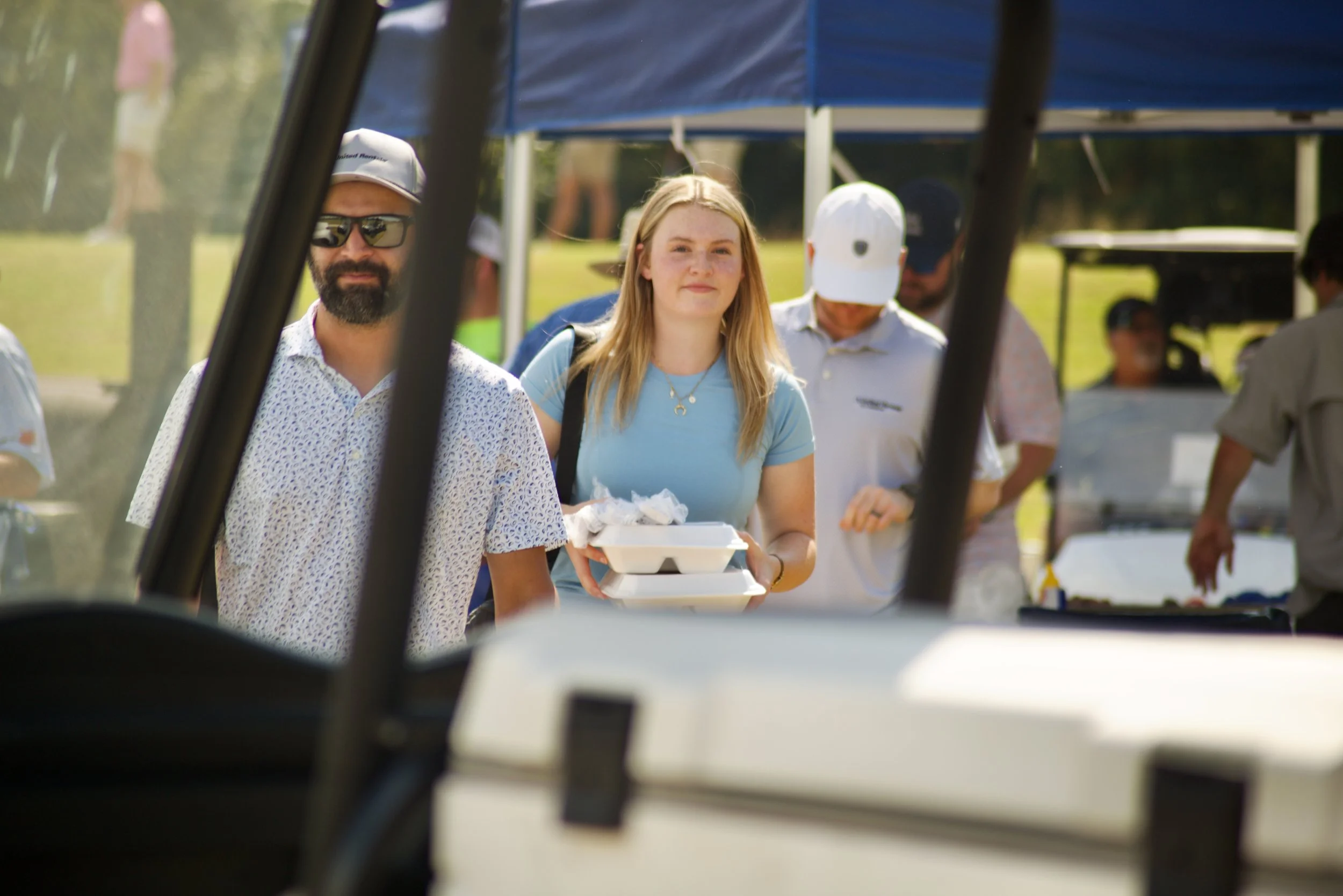 People in line at an outdoor food stall during daytime, with the photo taken from inside a golf cart.