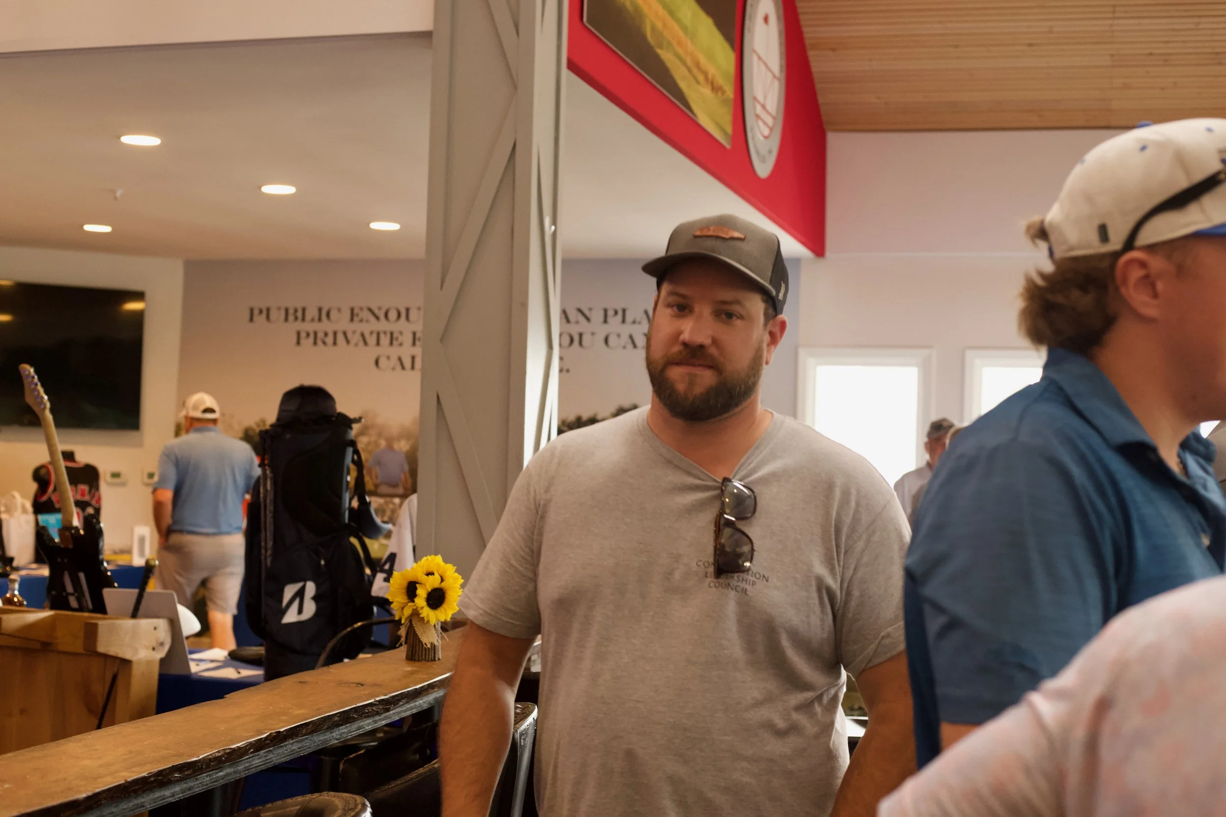 A man with a beard wearing a gray t-shirt and baseball cap stands in a busy indoor space, possibly a market or event, with other people in the background and a small sunflower bouquet on the table in front of him.
