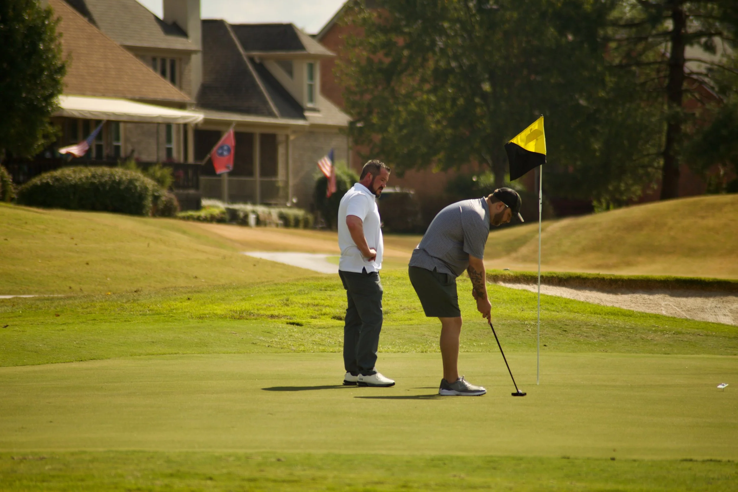 Two men playing golf on a green golf course with houses and trees in the background. One man is preparing to putt while the other is watching.