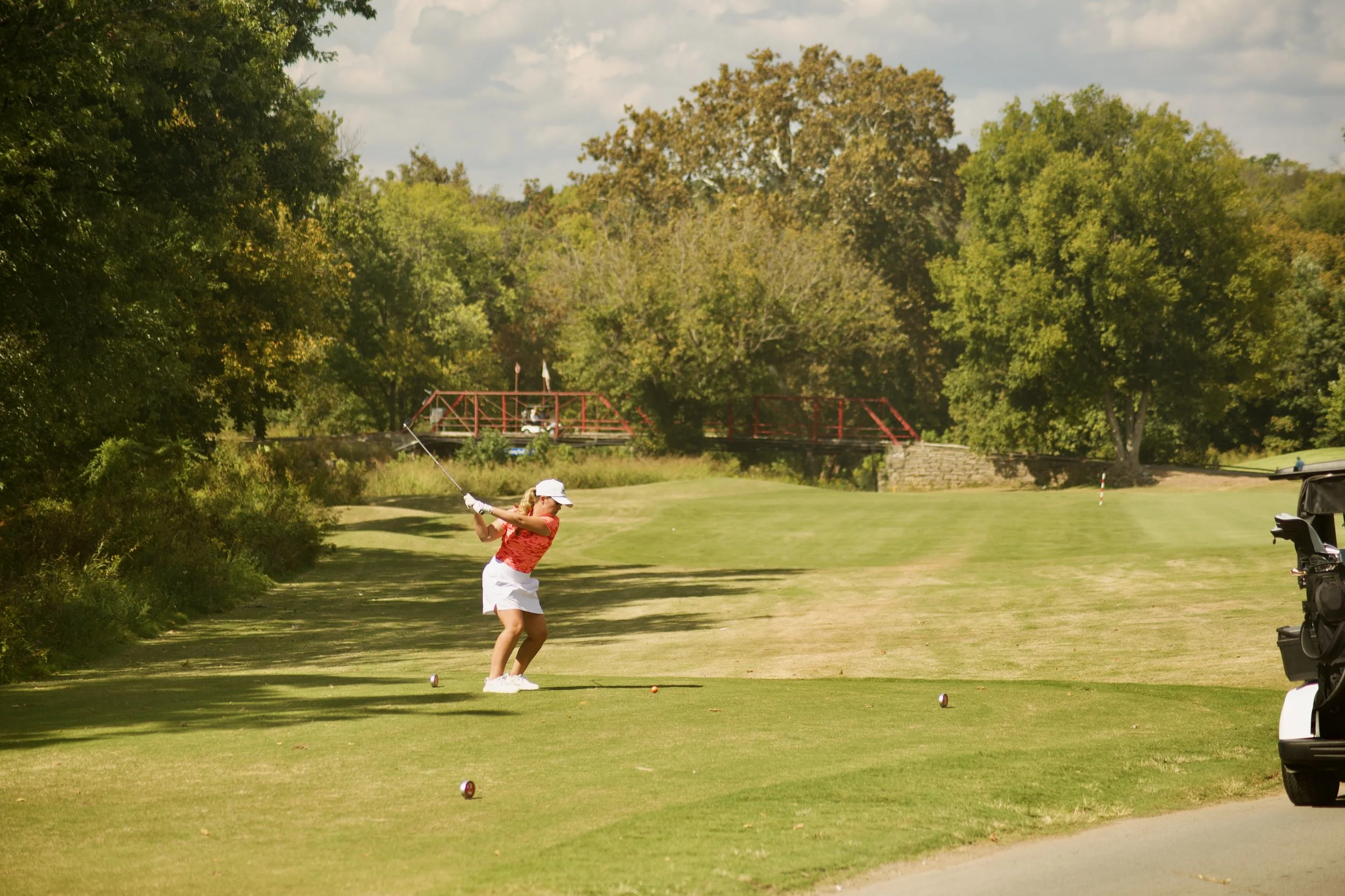 A woman in a red shirt and white skirt playing golf, preparing to swing, on a golf course with trees and a bridge in the background.