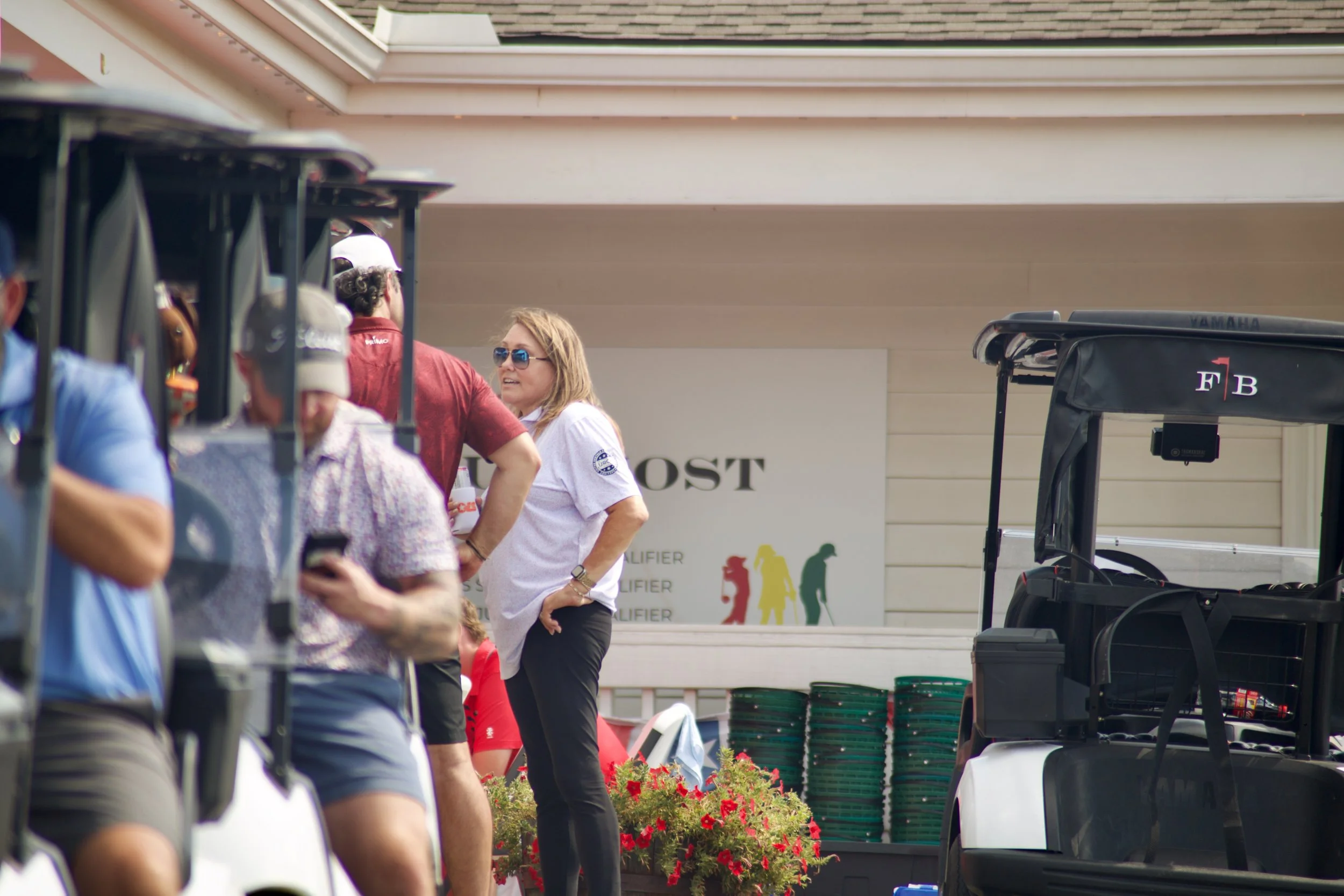 People standing near golf carts, some wearing sunglasses, with red flowers in the foreground and a sign with colorful silhouettes of people in the background.