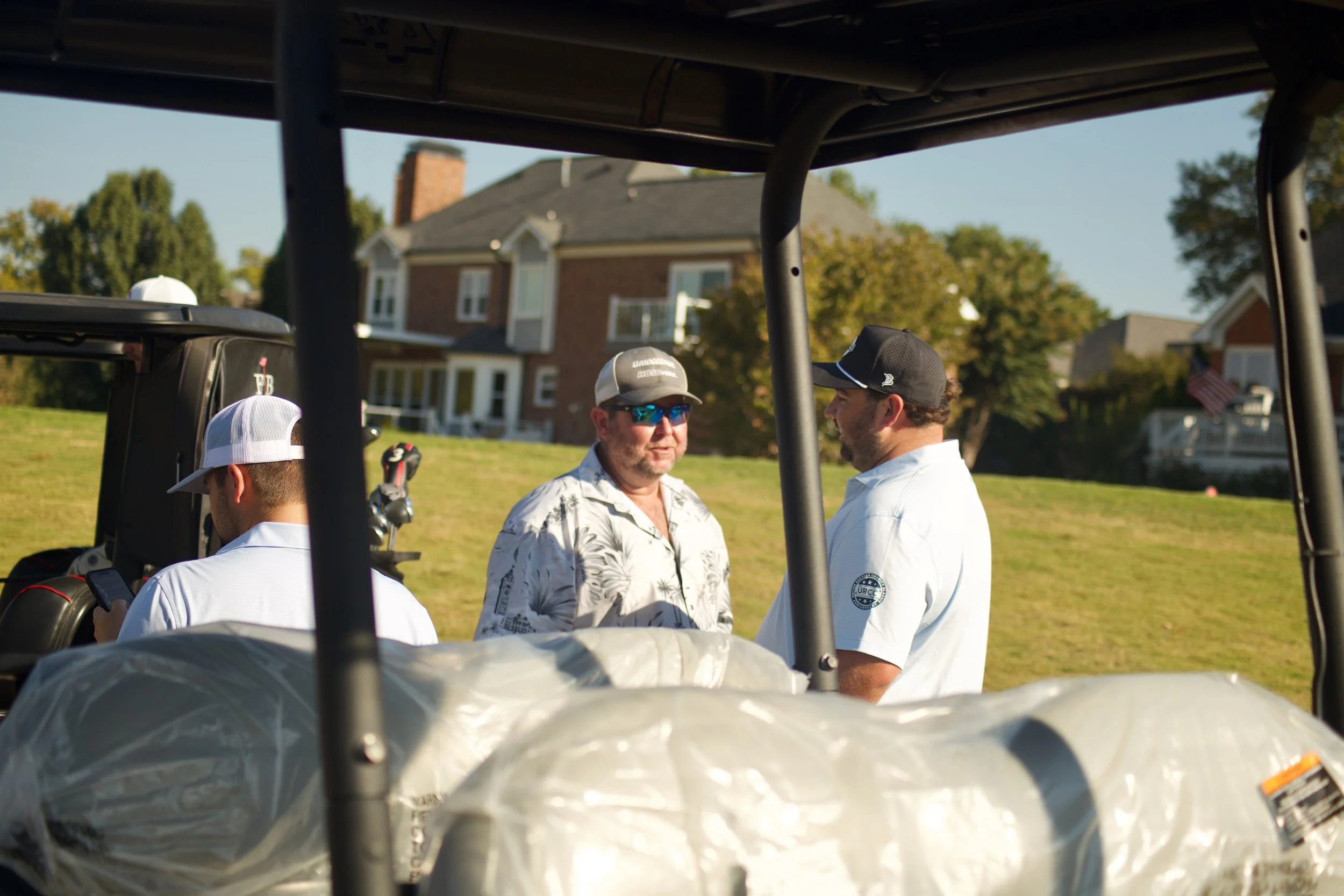 Three men standing outside near a house with trees, seen from inside a golf cart.