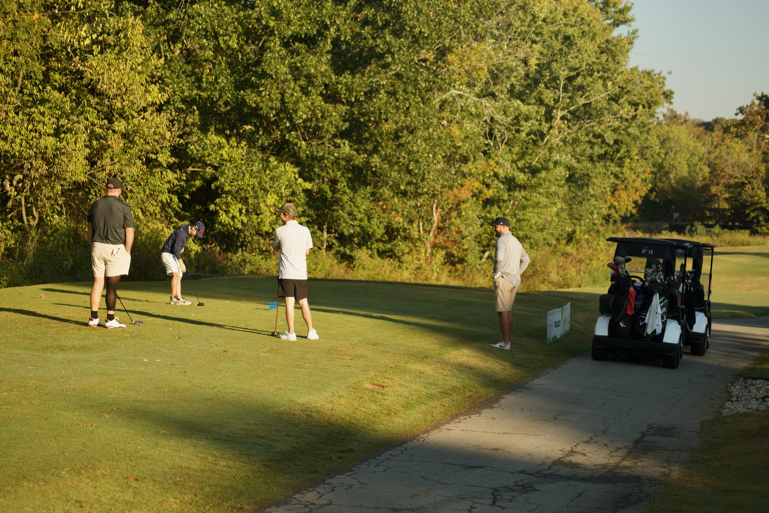 Four men playing golf on a green golf course with trees in the background and a golf cart parked nearby.