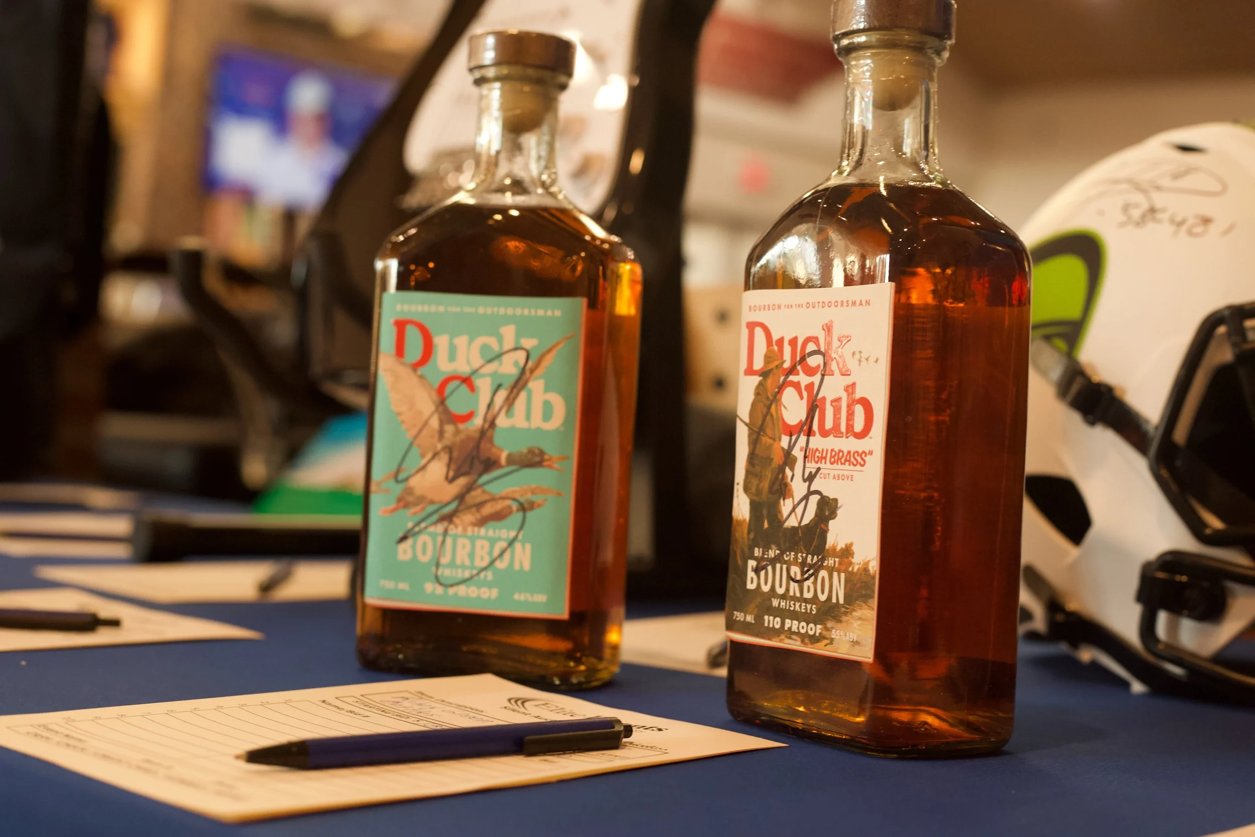 Two bottles of Duck Club bourbon whiskey on a table with a notepad and pen, with a blurred background showing a screen and other objects.